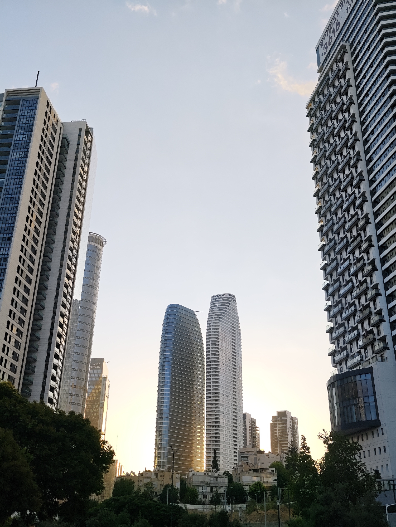 In Tel-Aviv, Israel, a low-angle shot captures a cluster of modern skyscrapers piercing a pale sky, illuminated by the soft glow of sunset. The buildings exhibit a variety of architectural styles, from the sleek, curved glass facades of two prominent towers in the center to the more angular, stepped design of the building on the far right, which features numerous balconies. Another tall, slender, cylindrical building with a patterned facade stands between the central curved towers and the building on the left. A particularly tall, rectangular skyscraper with a grid of windows and balconies dominates the left side of the frame, its peak slightly obscured by the sky. Lower structures and lush green trees fill the foreground, partially obscuring the bases of the skyscrapers and suggesting a transition from urban density to natural elements. The overall impression is one of contemporary urban development against a tranquil, natural backdrop.