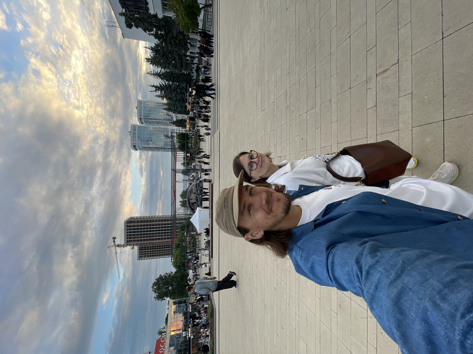 In an outdoor urban plaza, two individuals, a man and a woman, are captured in a selfie. The man, in the foreground on the left, wears a denim jacket over a white t-shirt and a tan baseball cap. He has shoulder-length brown hair and a beard. The woman, positioned to his right, wears a white hoodie, glasses, and carries a brown leather handbag. They are walking on a large, paved plaza with a pattern of light-colored rectangular stones.
In the background, a lively scene unfolds. Several people are walking and gathered, with tents and temporary structures suggesting an event. Behind them, a cityscape with several modern high-rise buildings under construction and completed is visible against a cloudy sky with patches of sunlight. A construction crane stands tall next to a partially built skyscraper. Trees and other buildings, including one with signage that appears to say "РОССИЯ" (Russia), are also present in the midground and background, indicating a developed urban environment. The overall impression is a public space with ongoing development and activity.