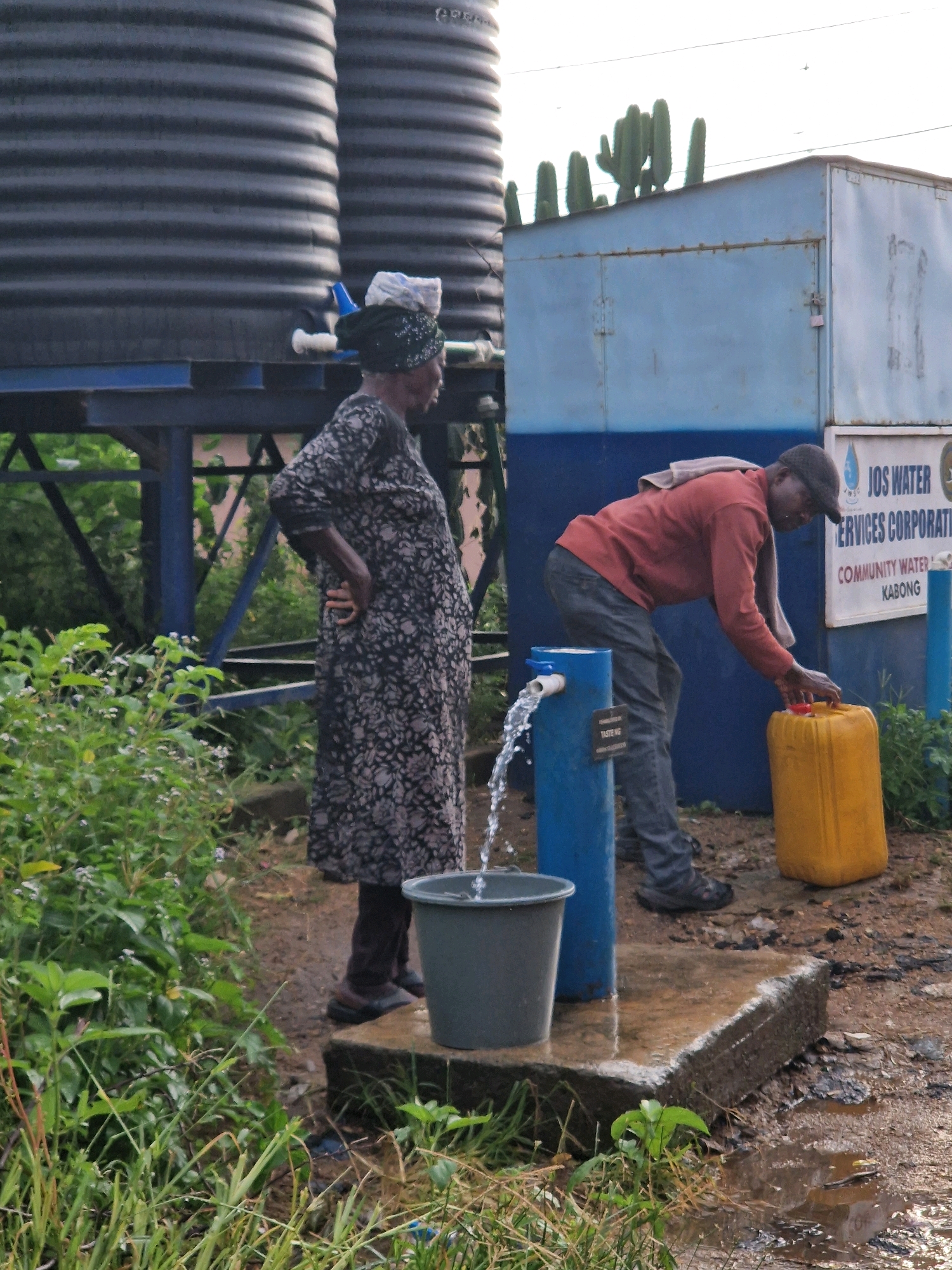 The image depicts an outdoor scene in Jos, Nigeria, where a man is filling a yellow jerrycan with water from a blue dispenser. The man, wearing a red long-sleeved shirt, dark trousers, and a dark baseball cap, is bent over, holding the jerrycan under the water spout. A light-colored cloth is draped over his shoulders. Behind him, a blue metal structure with a sign that reads "JOS WATER SERVICES CORPORATION COMMUNITY WATER KABONG" is visible. To the left of the dispenser, a woman in a dark floral-patterned dress and a headwrap stands with her hand on her hip, observing the scene. In the background, large black water tanks are mounted on a blue metal structure. The ground is muddy and wet, with some vegetation growing in the foreground.