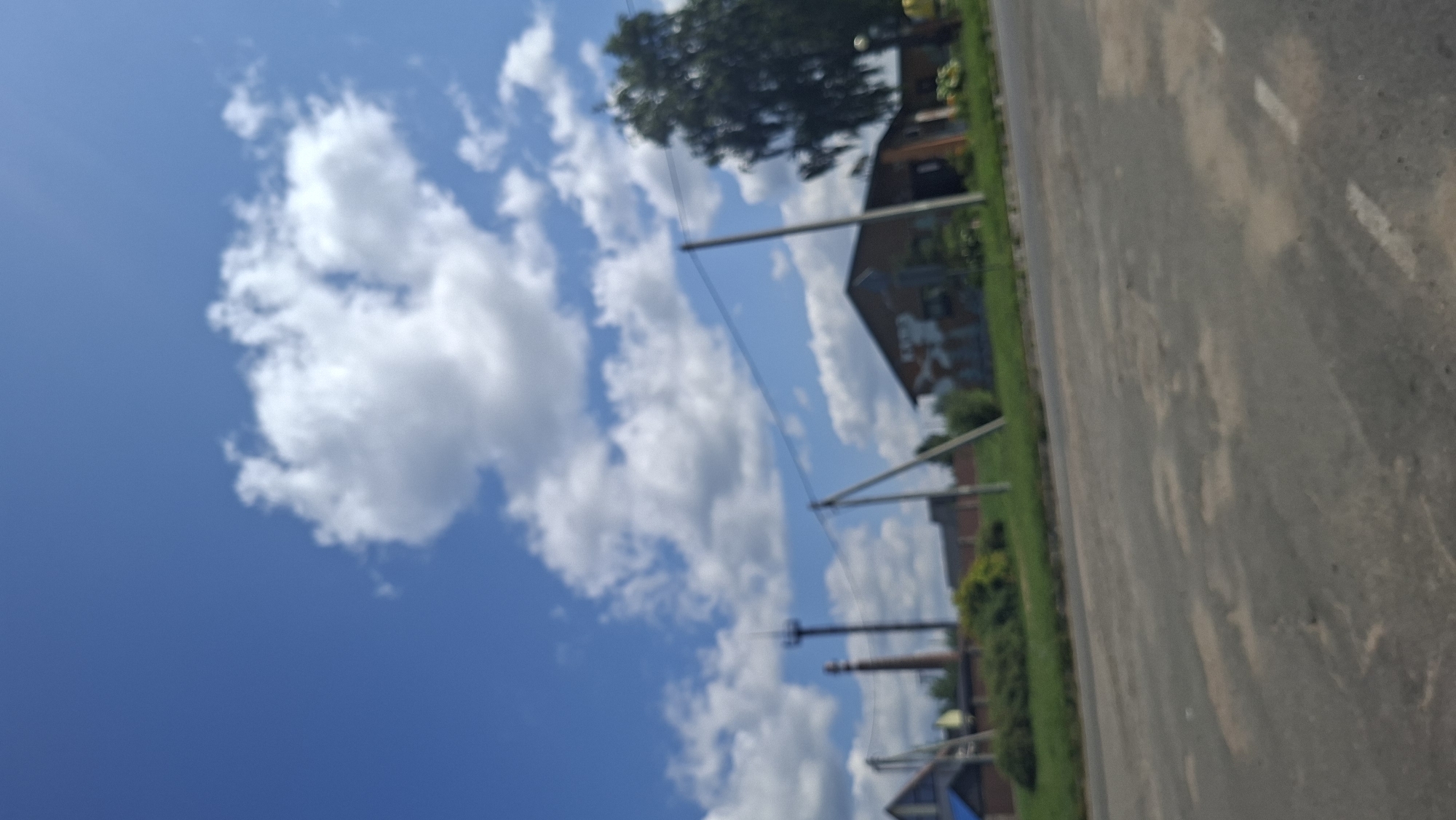 The image, taken from a low angle, shows a rural scene under a bright blue sky with scattered white clouds. In the foreground, a paved road with some light markings is visible. Beyond the road, a grassy area lines the roadside, with some bushes and trees. Several structures are present in the background, including a tall, slender tower with a structure at the top, possibly an industrial chimney or transmission tower. To the right of the tower, a building with a brown roof and light-colored walls, possibly a house or barn, is visible, with some graffiti-like markings on its exterior. Further to the right, a large, dark green tree with dense foliage stands prominently. Electrical wires are strung across the scene, supported by poles. The overall impression is of a residential or semi-industrial area on the outskirts of a settlement.
