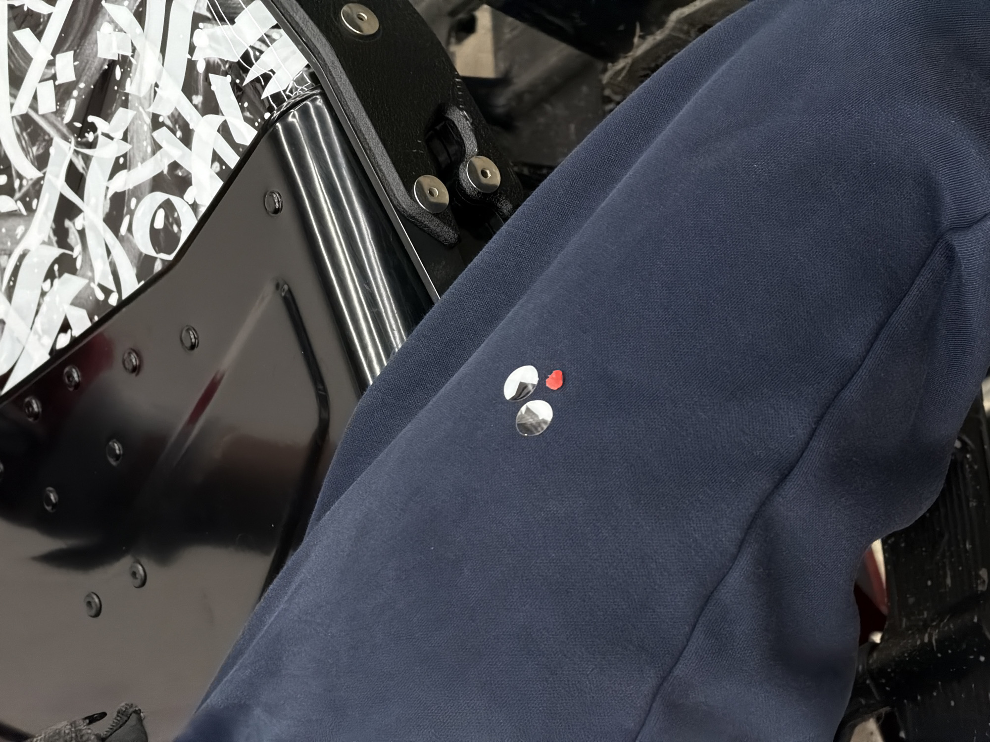 The image presents a close-up view of an interior setting. A portion of a leg, clad in dark blue fabric (possibly pants or a garment), occupies the right side of the frame. A small, shiny object with a red component is visible on the blue fabric.
To the left, a section of an unidentified object is visible. It features a black frame with silver rivets or fasteners. A portion of the object is covered in a white and black graphic design, possibly lettering or a stylized pattern. The object appears to be complex, featuring reflective surfaces and mechanical components.
The setting lacks specific details that would allow for an accurate determination of a location in Krasnoyarsk, Russia.