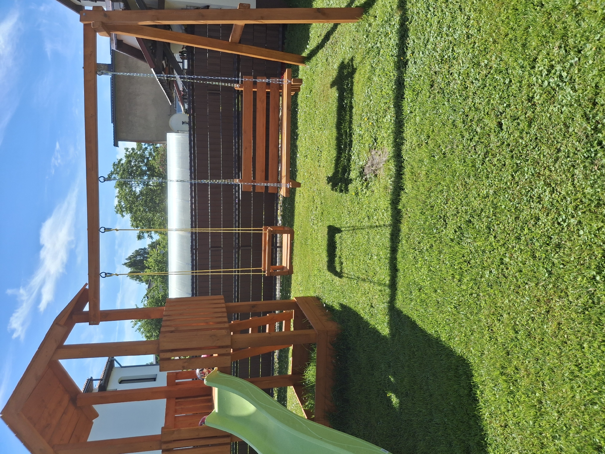 A wooden playground structure with a green slide, a single swing, and a bench swing is situated on a well-maintained lawn. The playground is constructed of light-colored wood and is positioned in a backyard with a brown wooden fence in the background. The sky above is clear blue with scattered white clouds. The scene is illuminated by bright daylight, casting distinct shadows from the playground equipment onto the grass. There are no people visible in the image. The location is a residential backyard in Ogre, Latvia.