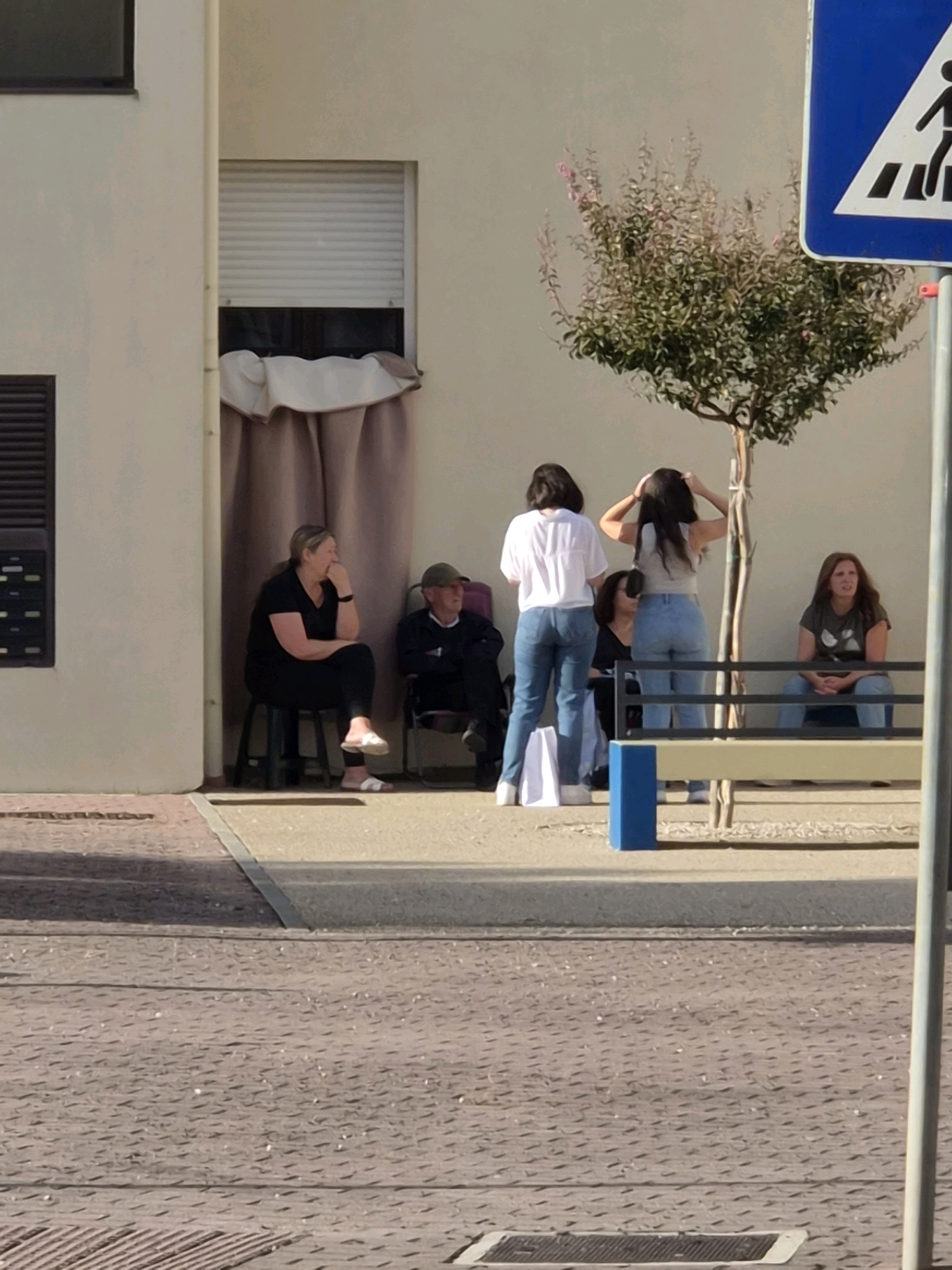 In Barcelos, Portugal, on a sunny day, five individuals are gathered outdoors near a building with a large window covered by a rolling shutter and a draped fabric. To the left, a woman in a black t-shirt and leggings sits on a black stool, with her hand on her chin. Next to her, a man in a dark jacket and green cap sits on a chair.
In the center of the scene, two young women stand facing away from the camera. One wears a white t-shirt and blue jeans, while the other wears a grey tank top and blue jeans, her hands adjusting her hair. A third woman sits on a bench to the right, wearing a green t-shirt and blue jeans, looking off to the side. A small tree with pink blossoms stands behind the women, and a blue and white pedestrian crossing sign is visible in the background. The ground in front of the building is paved with textured grey material.