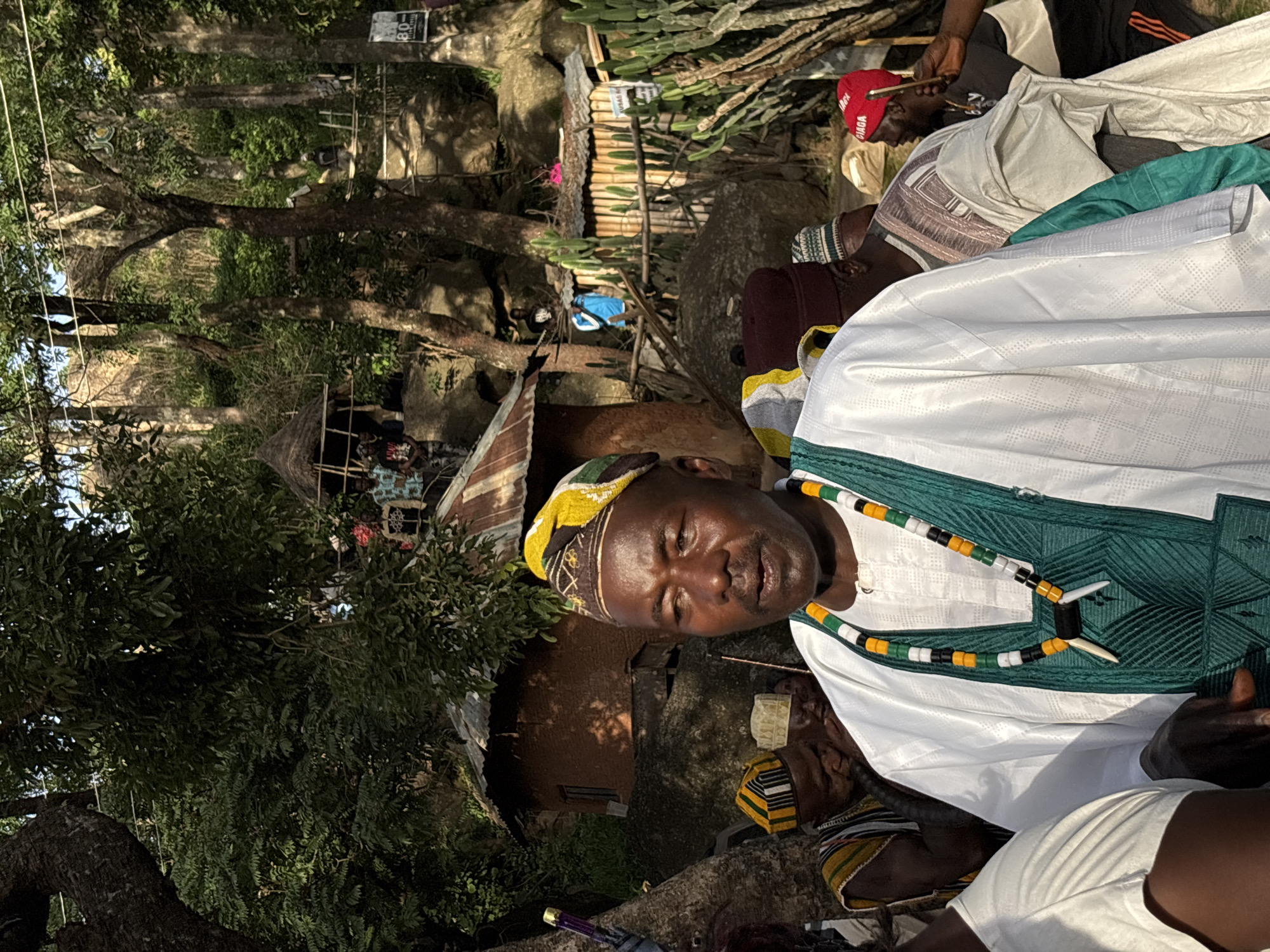 The image depicts a gathering of people, likely in a rural or semi-rural outdoor setting, possibly in Jos, Nigeria. The scene is bathed in natural light, suggesting it was taken during the daytime, perhaps late afternoon given the warm tones.
The central figure is a man wearing a white robe with intricate green embroidery around the neckline and chest. He has a colorful beaded necklace around his neck, featuring black, white, green, and yellow beads, and a yellow and green patterned headwrap. His expression is serious, and he is looking directly at the camera.
Other individuals are visible in the frame, some wearing traditional attire, including caps and robes. One person is wearing a red cap with white lettering. The background reveals a rustic environment with natural elements like large rocks and trees, along with some rudimentary structures that appear to be huts or sheds with thatched roofs and corrugated metal. There is also vegetation, including what appears to be a cactus plant.
The overall atmosphere seems to be one of community or a communal event. The presence of multiple people suggests a social gathering, a religious ceremony, or a traditional function. The ground is dirt, and the general impression is of a place that is not highly developed. There is a sign with some visible text, including "80-" and what appears to be "NO", but the full context is unclear. The image is oriented vertically, with the subjects appearing sideways.