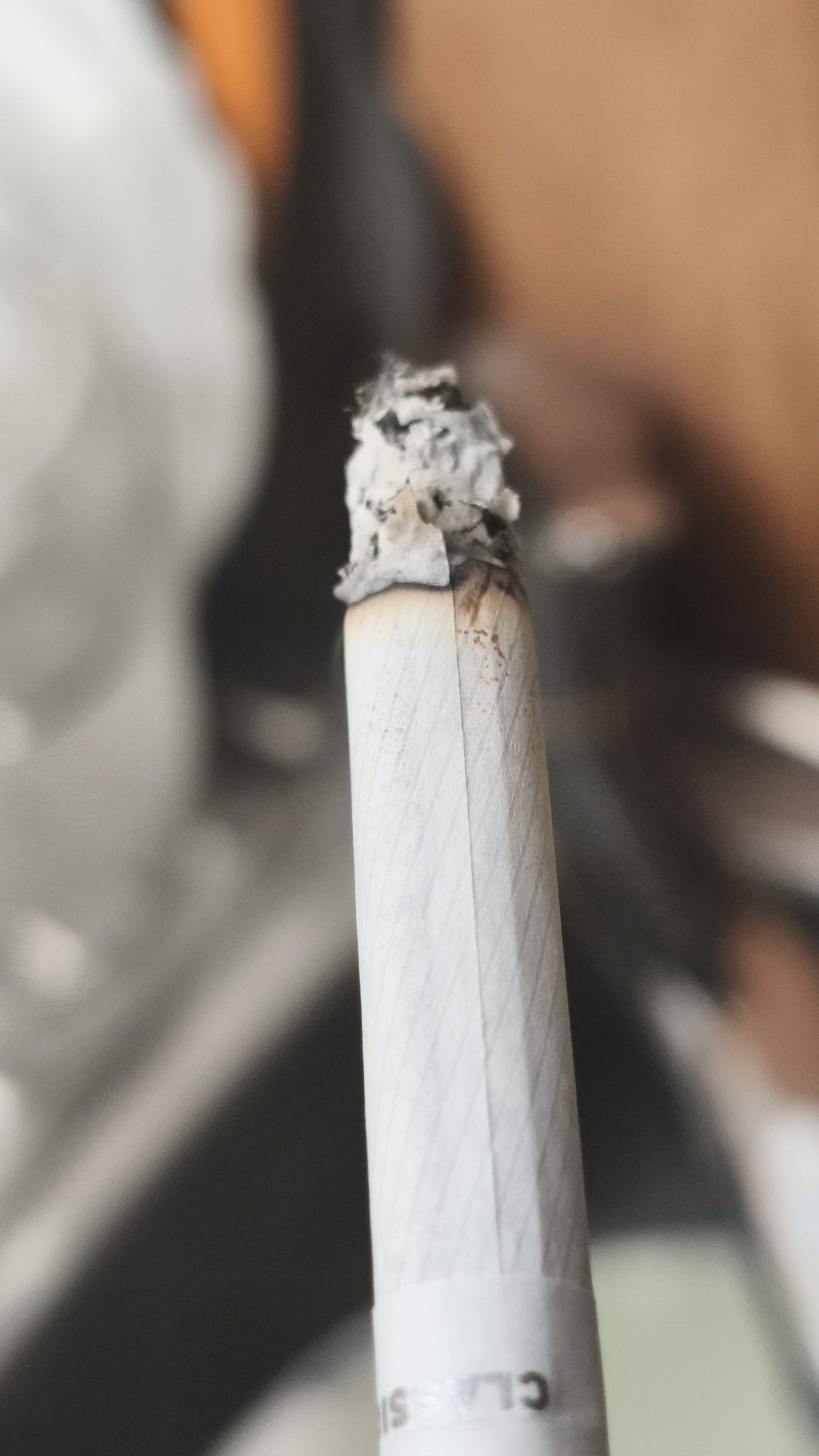 A close-up shows a lit cigarette with a growing ash tip. The cigarette is white with a textured paper covering, and the partial word "CLASSI" is visible at the bottom. The background is blurry and indistinct. There is no indication of this being Nowy Tomyśl, Poland.