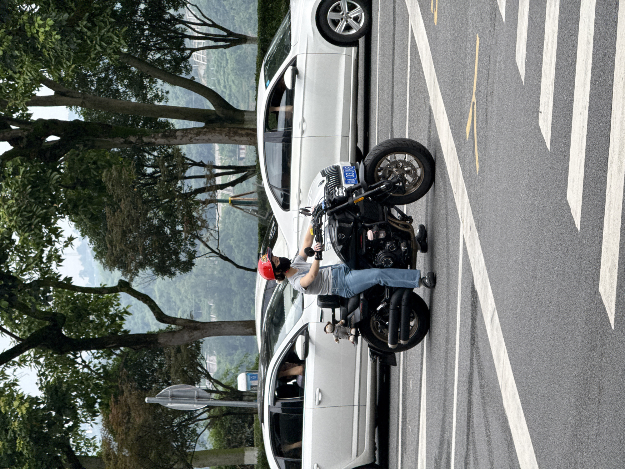 A medium shot shows a woman on a black motorcycle stopped at a crosswalk on a city street in Jiangjin, China. She is wearing a red helmet, a grey t-shirt, and blue jeans. The motorcycle has a plush toy attached to its rear. Two white cars are visible behind the motorcycle. The street is paved with asphalt, marked with white lines. Trees and hills form the background. The sky is overcast.