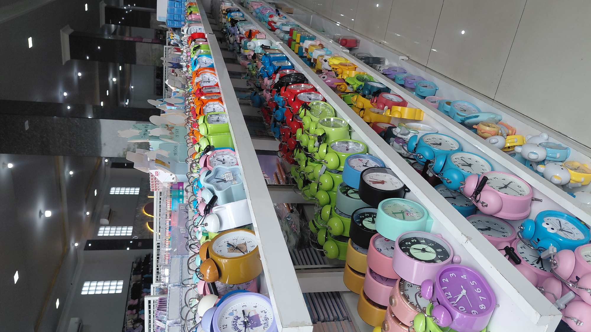This is a medium shot, eye-level, indoor photograph of a retail store display filled with numerous alarm clocks. The clocks are arranged on white shelves that extend from the left foreground to the right background of the image. The shelves are packed with alarm clocks in a wide variety of colors, including pink, purple, blue, green, yellow, orange, red, and white. Many of the clocks are classic twin-bell alarm clock designs, while others have more modern or novelty shapes. Some clocks feature cartoon characters or designs on their faces. In the background, more merchandise is visible, including stationery items like colored pencils and notebooks, as well as what appear to be other display shelves and store fixtures. The store appears to be well-lit with fluorescent ceiling lights. The overall impression is of a brightly stocked gift shop or stationery store. Given the context of Purwokerto, Indonesia, this is likely a section of a department store or a dedicated gift shop. There are no people visible in the image.