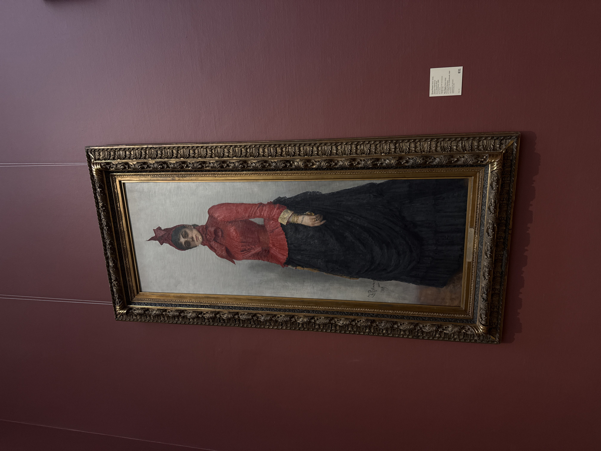 A framed portrait of a woman hangs on a burgundy wall. The portrait has a gold frame. The woman in the painting is wearing a red dress and a black skirt. A small white sign is affixed to the wall above the painting. The image was taken in Moscow, Russia.