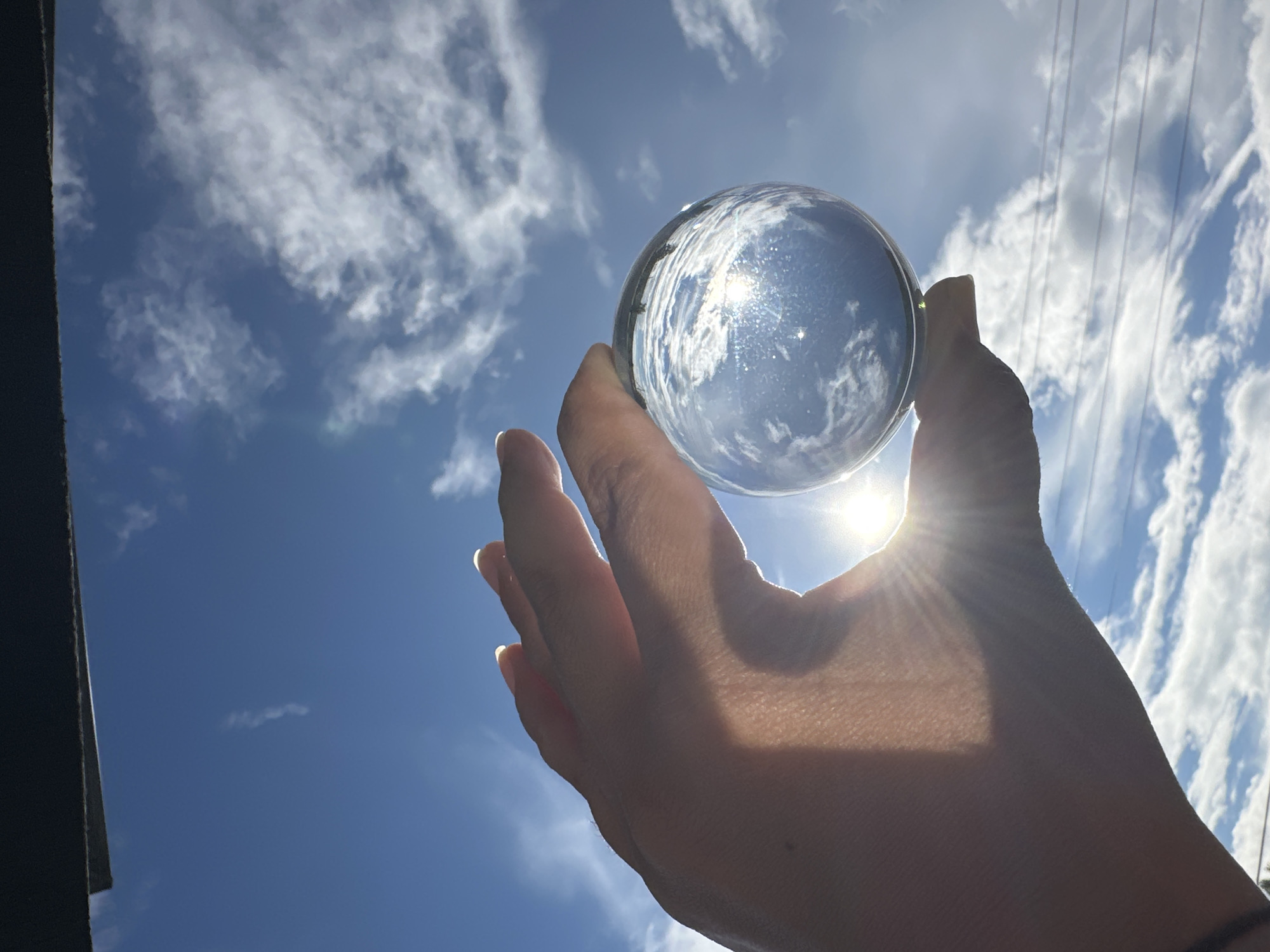 A human hand, silhouetted by intense sunlight, firmly grasps a clear spherical object against a vibrant blue sky filled with scattered white cumulus clouds. The sun, positioned behind the sphere and hand, generates a prominent sunburst effect and lens flares, both in the direct sky and magnified within the sphere itself, which reflects an inverted image of the surrounding sky. The hand, illuminated from behind, shows skin texture and well-defined fingernails; a dark band or bracelet is visible on the wrist. A dark, solid horizontal structure, likely a roofline or overhang, spans the top edge of the frame. In the lower right background, several thin, dark horizontal lines, consistent with utility wires, are visible against the sky. The scene is outdoors, under bright daylight conditions, in the city of Unknown, Unknown.