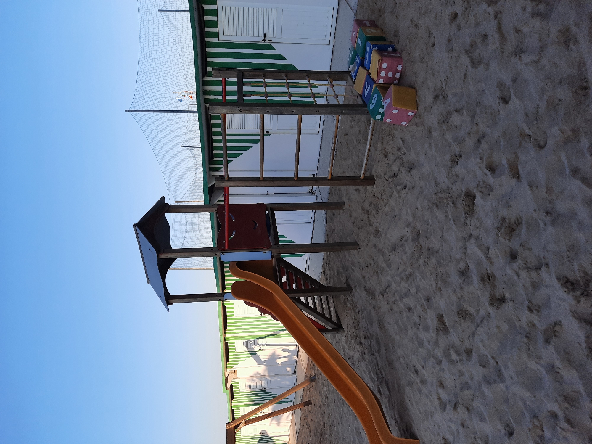 The image depicts a children's playground on a sandy beach, likely in Pesaro, Italy, given the context. In the foreground, the sand is textured with footprints and slight undulations. To the left, an orange slide descends from a wooden play structure. This structure features a small, dark-roofed tower with a red smiley face panel and a set of wooden stairs. Adjacent to the slide is a wooden swing set with two angled support beams.
To the right of the play structure, a set of wooden monkey bars is visible, along with a rope net climbing element. In front of this, a row of colorful, oversized dice-shaped cushions are arranged on the sand. These cushions are primarily pink, blue, yellow, and green, and display large white numbers and dots, resembling dice.
In the background, a row of beach cabins with alternating green and white striped facades stretches across the scene. These cabins have white doors and shutters, and their shadows are cast on the sand. Above the cabins, a fine mesh net, possibly a safety net for a sports court, is suspended between wooden poles, with a clear blue sky above. The overall scene suggests a public recreational area on a beach, likely during daylight hours, given the bright sky and visible shadows.