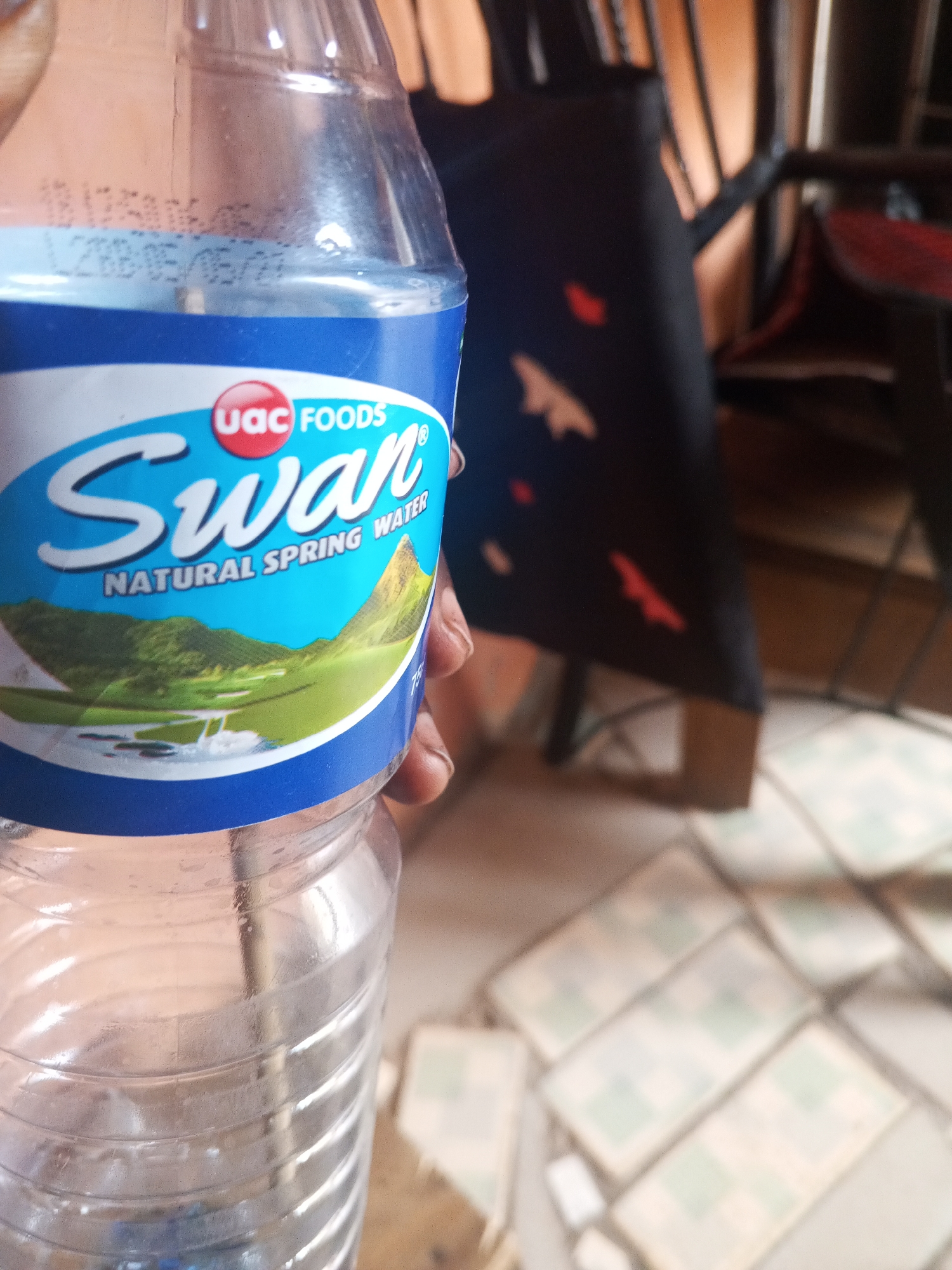 A plastic bottle of Swan Natural Spring Water produced by UAC Foods is held in someone's hand. A date code is printed on the bottle: "L288-05/05/22". The bottle is partially empty. In the background, there's a black and red patterned fabric draped over a chair, and tiled floor is visible. The location is Gwong, Nigeria.