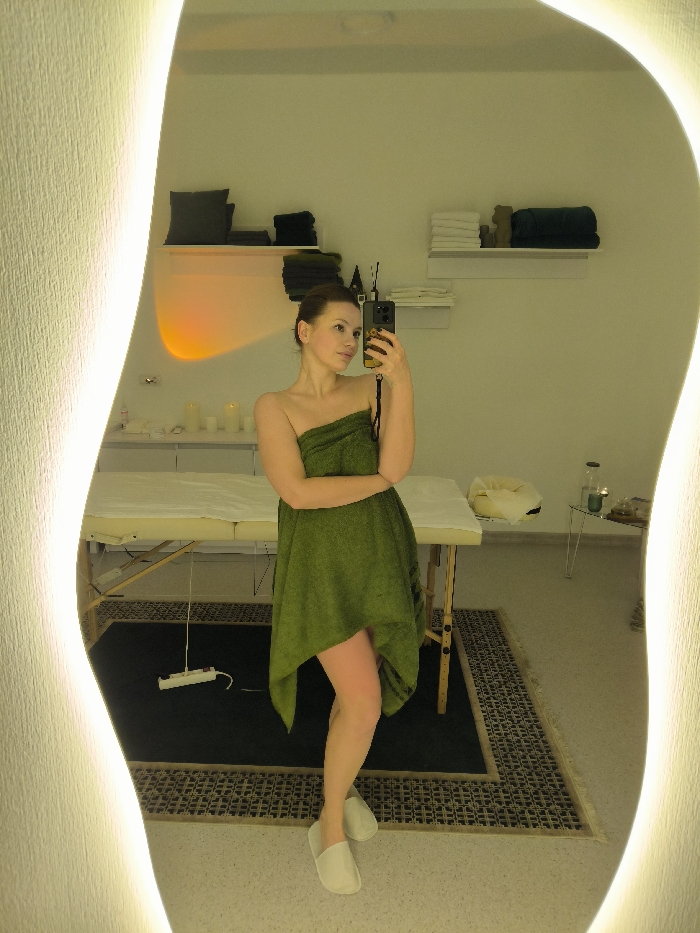 A female subject is captured in a mirror selfie within what appears to be a spa or treatment room in Vinnytsia, Ukraine. The subject, with light brown hair pulled back, is wrapped in a dark green towel that covers her torso and extends asymmetrically, reaching approximately mid-thigh on one side and revealing more of the other leg. She wears light-colored disposable slippers and holds a dark-colored smartphone with a patterned case in her right hand, positioned to take the photograph.
The room's interior, reflected in the large, irregularly shaped mirror with illuminated, wavy edges, features light-colored walls and a floor with a patterned dark rug beneath a light-colored massage table covered with a white sheet. To the left of the massage table, a white counter holds small white objects, and an orange light glow emanates from the wall above it. Above this counter, two white floating shelves display dark cushions and folded dark towels. To the right of the massage table, a small, minimalist metal-framed table holds a clear bottle, small containers, and a light-colored fabric container. Two additional white floating shelves above this table display stacks of folded white and dark towels. An off-white power strip with a white cord is visible on the floor near the massage table's left leg. The overall lighting is bright and warm, accented by the mirror's yellow backlighting.