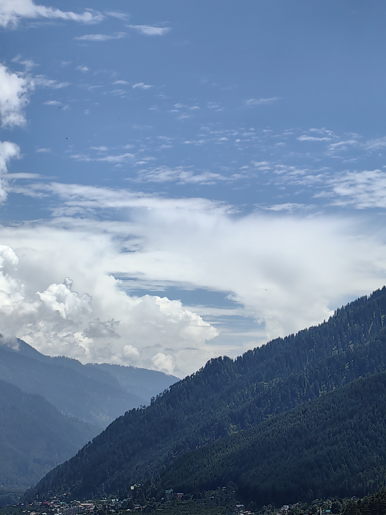 The image displays a panoramic view of a mountainous landscape, likely in or around Vashisht, India, characterized by lush green slopes covered in dense pine trees. In the foreground, a village with numerous buildings is visible nestled at the base of the mountains. The sky is a vibrant blue, punctuated by scattered white clouds, some of which are large and cumulus, while others are thin and wispy. The overall scene evokes a sense of natural beauty and a tranquil rural setting.