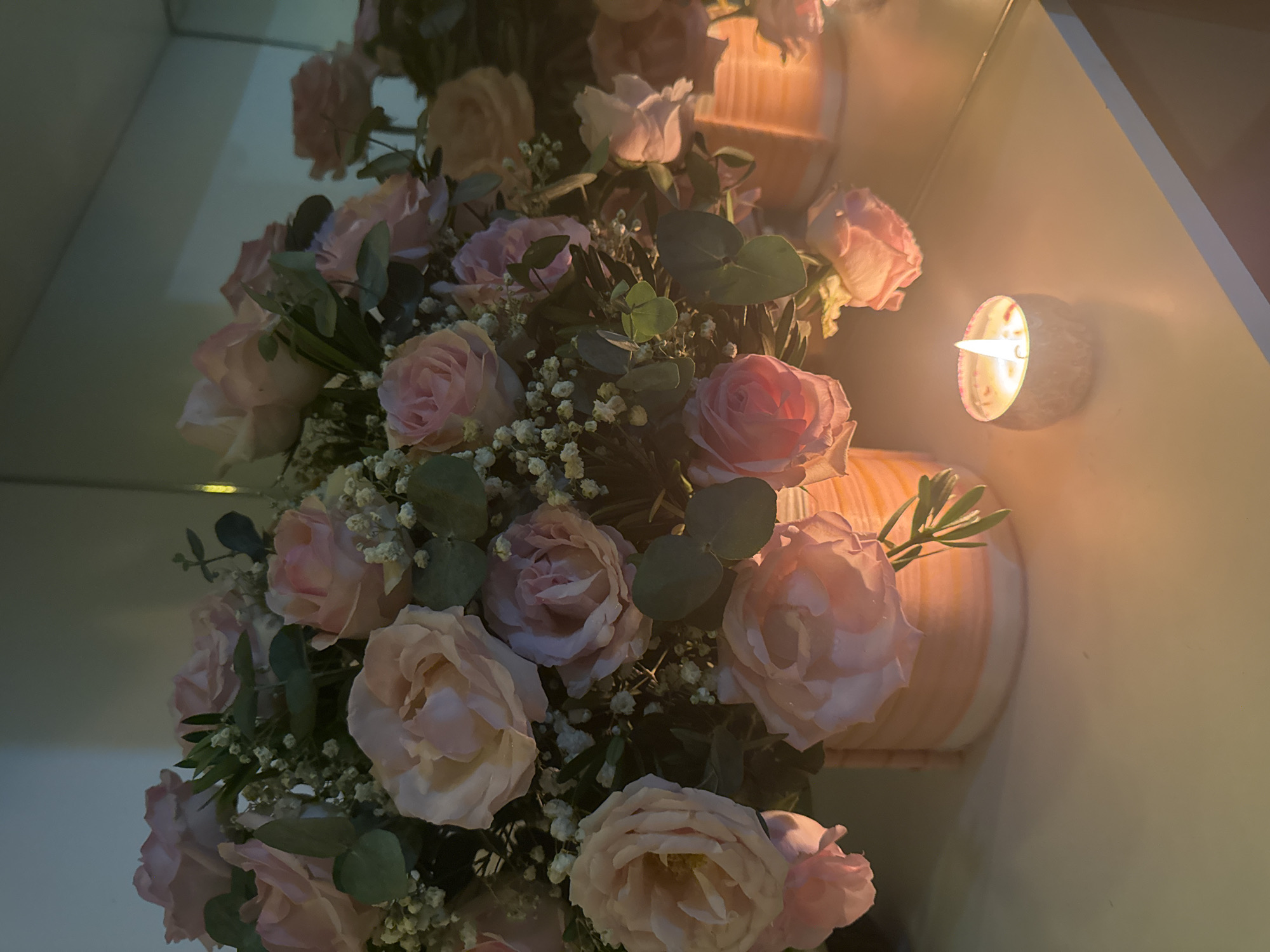 The image presents a close-up, angled view of a beautiful floral arrangement and a lit candle, set in an indoor environment. The main subject is a lush bouquet dominated by numerous light pink roses, some fully open and others still in bud. These roses are accented with delicate sprigs of white baby's breath (gypsophila) and clusters of green, round-leafed foliage, possibly eucalyptus. The flowers exhibit soft, delicate petals and appear fresh.
To the right of the floral arrangement, a single tea light candle glows brightly within a small, textured, cylindrical holder. The candle's flame casts a warm, orange-yellow light that illuminates the edges of the flowers and the surrounding surfaces, creating a soft and inviting ambiance. The bouquet itself is situated in a light-colored, possibly striped or ribbed, cylindrical vase or container, which is partially visible on the right side of the frame.
The background features light-toned walls, with a distinct corner or reflective surface (potentially a mirror) visible on the upper left, showing blurred reflections of the flowers and surrounding elements. The dim overall lighting, accentuated by the candle's glow, suggests the scene is set during the evening or night. The atmosphere conveyed is one of warmth, tranquility, and elegance. No text is visible in the image.