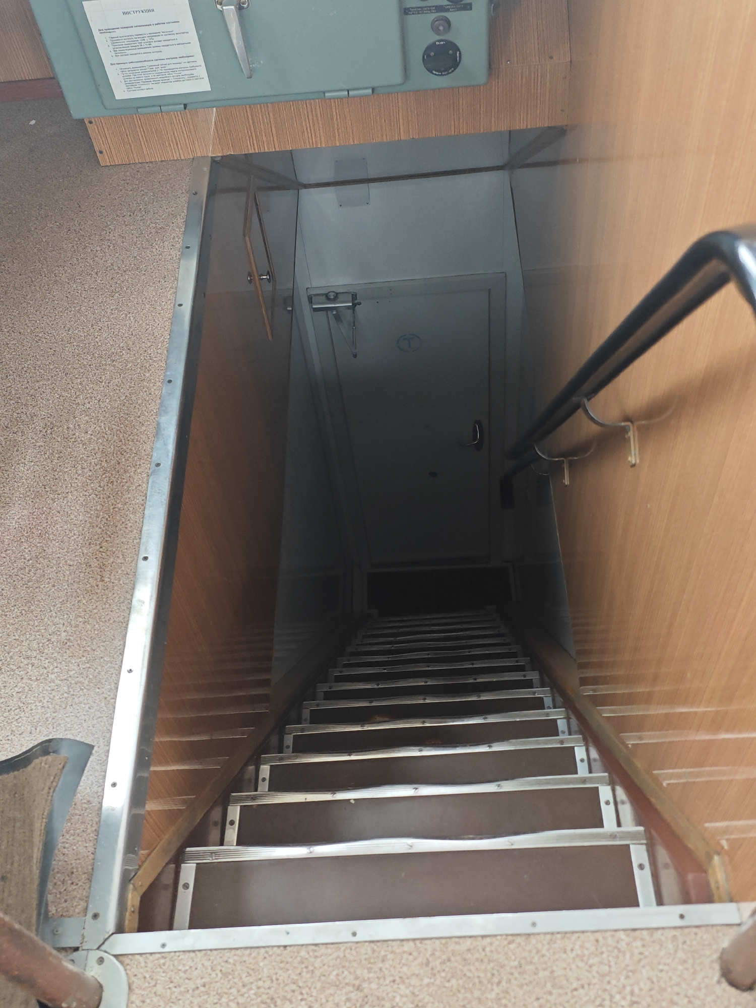 The image shows a downward view of a dimly lit staircase. The staircase has wooden steps with metal trim and a metal handrail on the right side. The walls on either side appear to be made of wood paneling and carpeted surfaces. At the top of the stairs, there is a door with a handle and a small circular symbol. Above the door, a metal panel with some text and a lever is visible. The scene appears to be in an interior space, possibly a building or a vehicle, and there are no people visible. The location of Krasnoyarsk, Russia is not evident from the image itself.