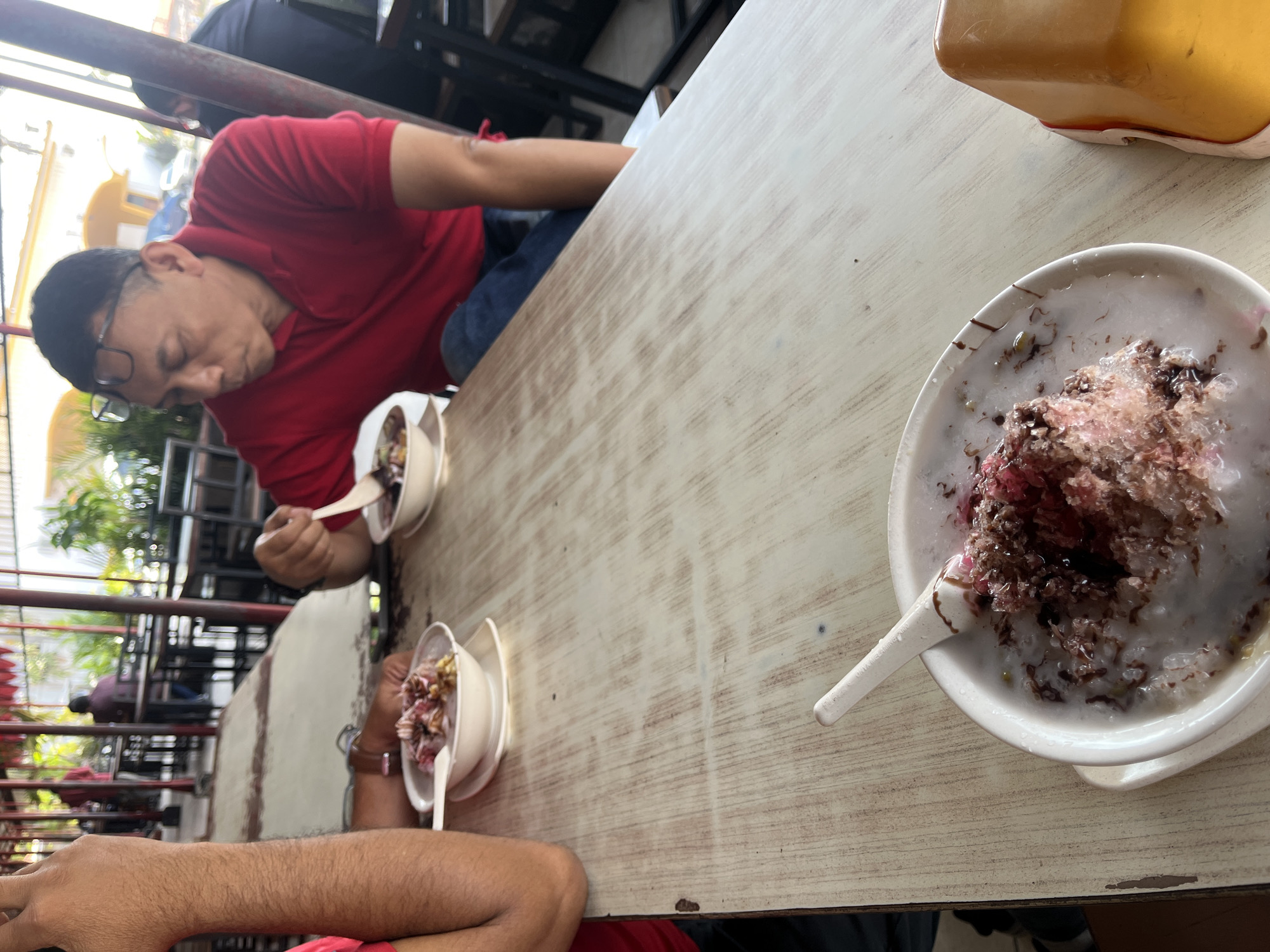 The image captures a close-up view of a man in a red polo shirt sitting at a wooden table, likely in an outdoor or semi-outdoor dining area, possibly a warung or a restaurant in Singosari, Indonesia. He is wearing glasses and appears to be eating a dessert from a white bowl with a spoon. The dessert is a shaved ice concoction with toppings that include red syrup, what looks like green beans or jelly, and possibly condensed milk. In the foreground, another bowl of dessert is partially visible, also with a spoon. The table has a light-colored, weathered wood finish. In the background, other diners and a person in uniform are visible, along with structural elements like red pillars and a roof, suggesting a busy food establishment. The overall atmosphere seems casual and lively.