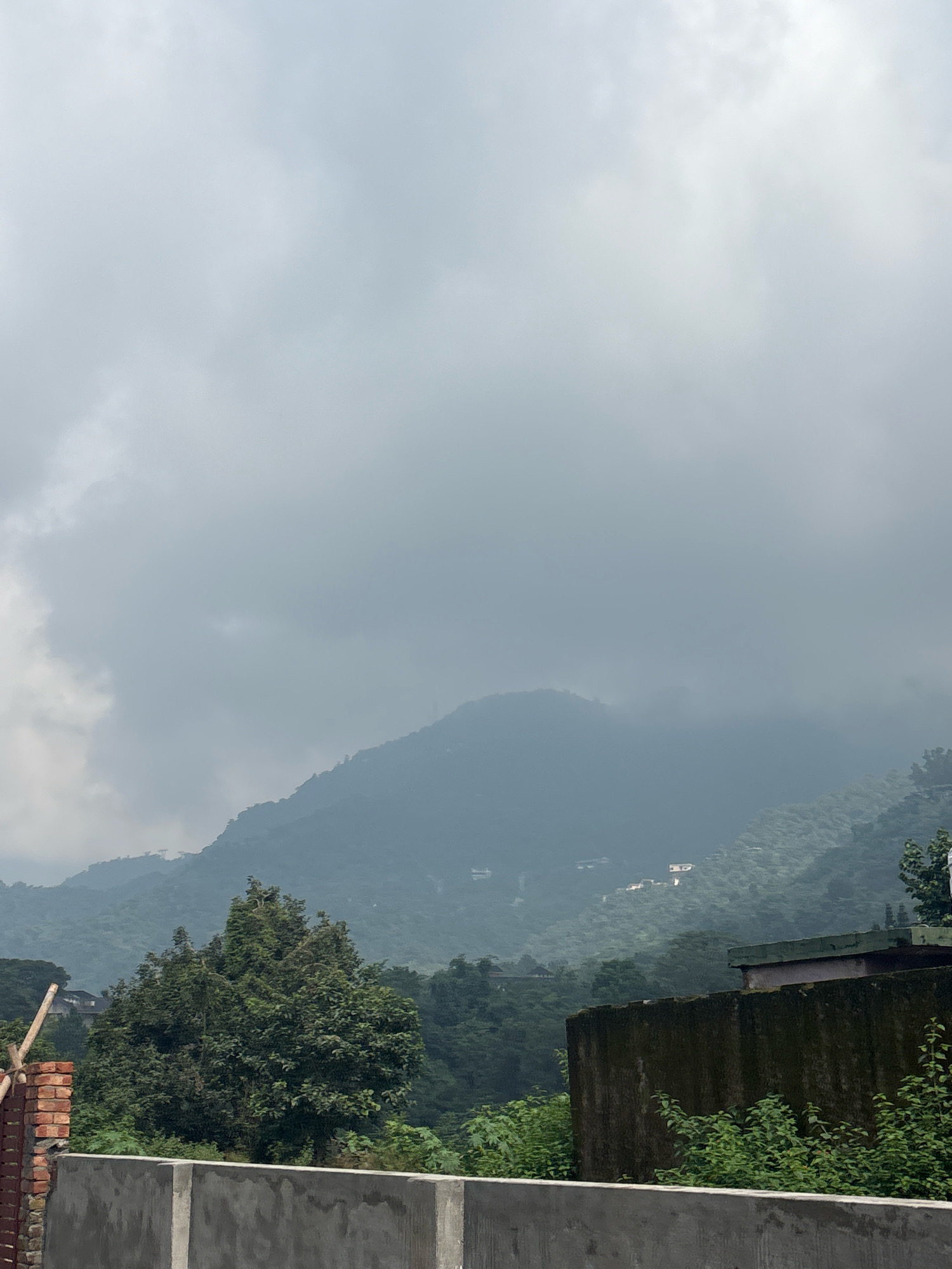 The image depicts a hazy, overcast sky over a lush, mountainous landscape. In the foreground, a concrete barrier runs horizontally across the bottom of the frame, with some greenery and a brick structure on the left side. Beyond the barrier, dense foliage and trees dominate the mid-ground. Several buildings are visible scattered across the slopes of the mountains in the background. The atmosphere is diffused, suggesting a misty or rainy day, common in the hilly regions of Bhatta, India.