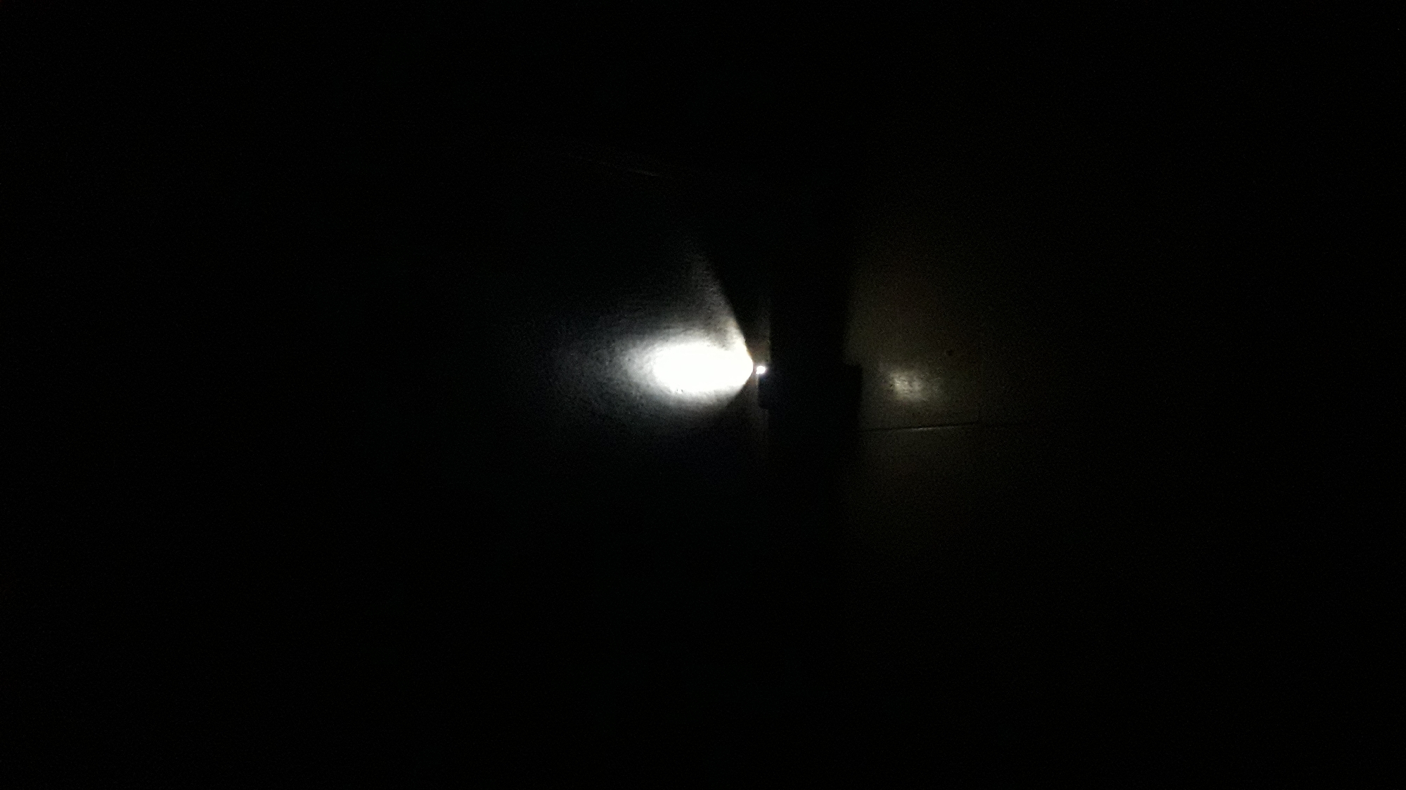 The image is a low-light, close-up shot of a small, dark-colored object emitting a bright white light. The object appears to be rectangular or square and is positioned on a dark, textured surface that resembles a tiled floor. The light from the object casts a bright, diffused glow on the surface in front of it and illuminates a rough, textured wall behind it, creating a halo effect. The surrounding environment is predominantly black, indicating a dark or unlit setting, likely indoors. There are no people or discernible interactions visible in the image. The scene appears to be a simple illumination of a surface and wall by a light source, with no context of its specific location within Nkpor, Nigeria, or any other contextual elements.
