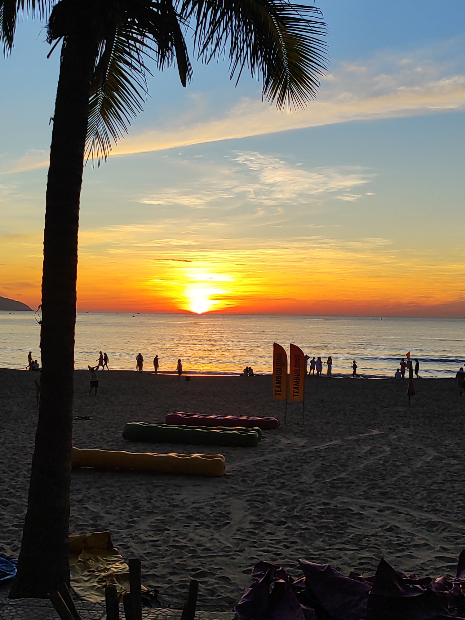 A silhouette of a palm tree is in the foreground, its fronds reaching towards a sky painted with the warm hues of sunrise over the sea. The ocean is calm, reflecting the vibrant orange and yellow gradient of the sky, transitioning into a soft blue above. The beach is dotted with several people, some walking along the shoreline, others gathered in small groups. Two orange flags with the text "TEAMBUILDING" are planted in the sand. In the mid-ground, there are several colorful, inflatable, kayak-like objects lying on the sand. The sand itself is textured, showing ripples and footprints. The overall atmosphere suggests a peaceful morning scene at a beach in Da Nang, Vietnam, possibly during a team-building event.