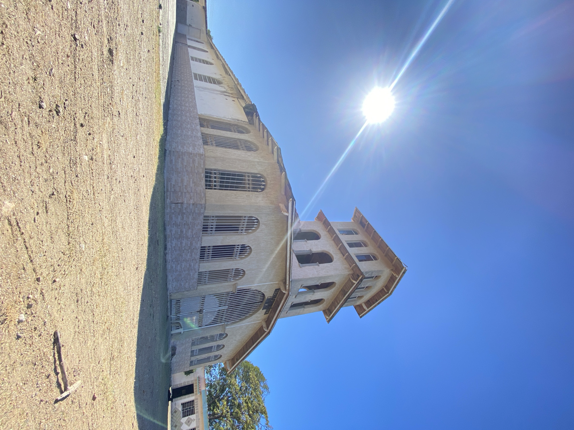 The image shows the Iglesia de San Juan Evangelista in Juan Griego, Venezuela, taken from a low angle. The church has a light-colored facade with arched windows protected by metal bars, and a tiered tower. The sky is a clear blue with a bright sun positioned in the upper right. A portion of the ground, covered with dry vegetation and dirt, is visible on the left. The composition is vertical.