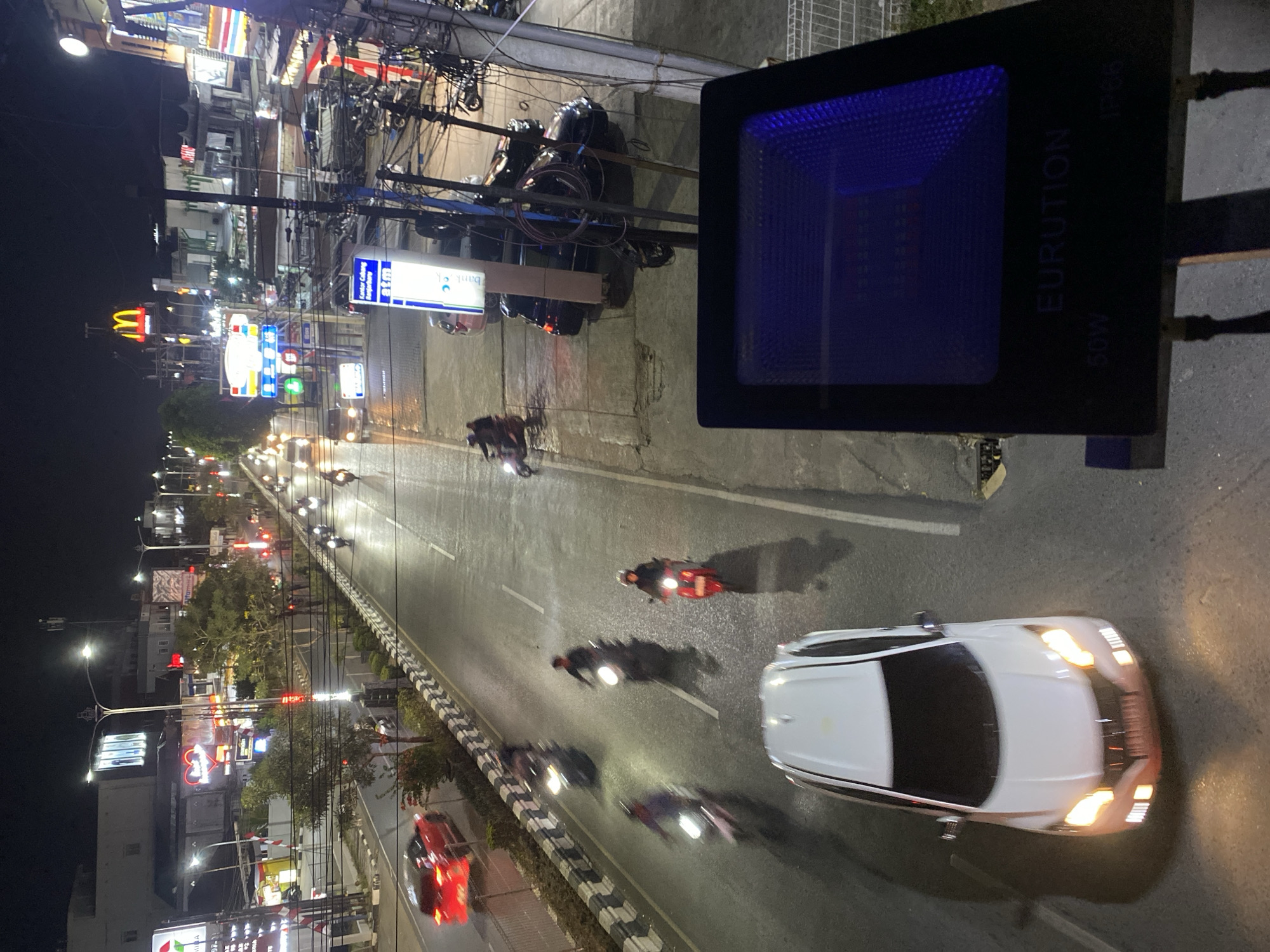 The image shows a nighttime street scene in Banjarbaru, Indonesia. A multi-lane road is visible, with several vehicles, including cars and motorcycles, moving in both directions. The road is illuminated by streetlights and the headlights and taillights of the vehicles. Buildings with illuminated signs are present on either side of the road, suggesting a commercial or urban area. A McDonald's sign is prominent on the right side of the image. In the foreground, a floodlight with the brand "EURUTION" is visible, partially obscuring the view of the street. The overall impression is of a busy urban environment at night.