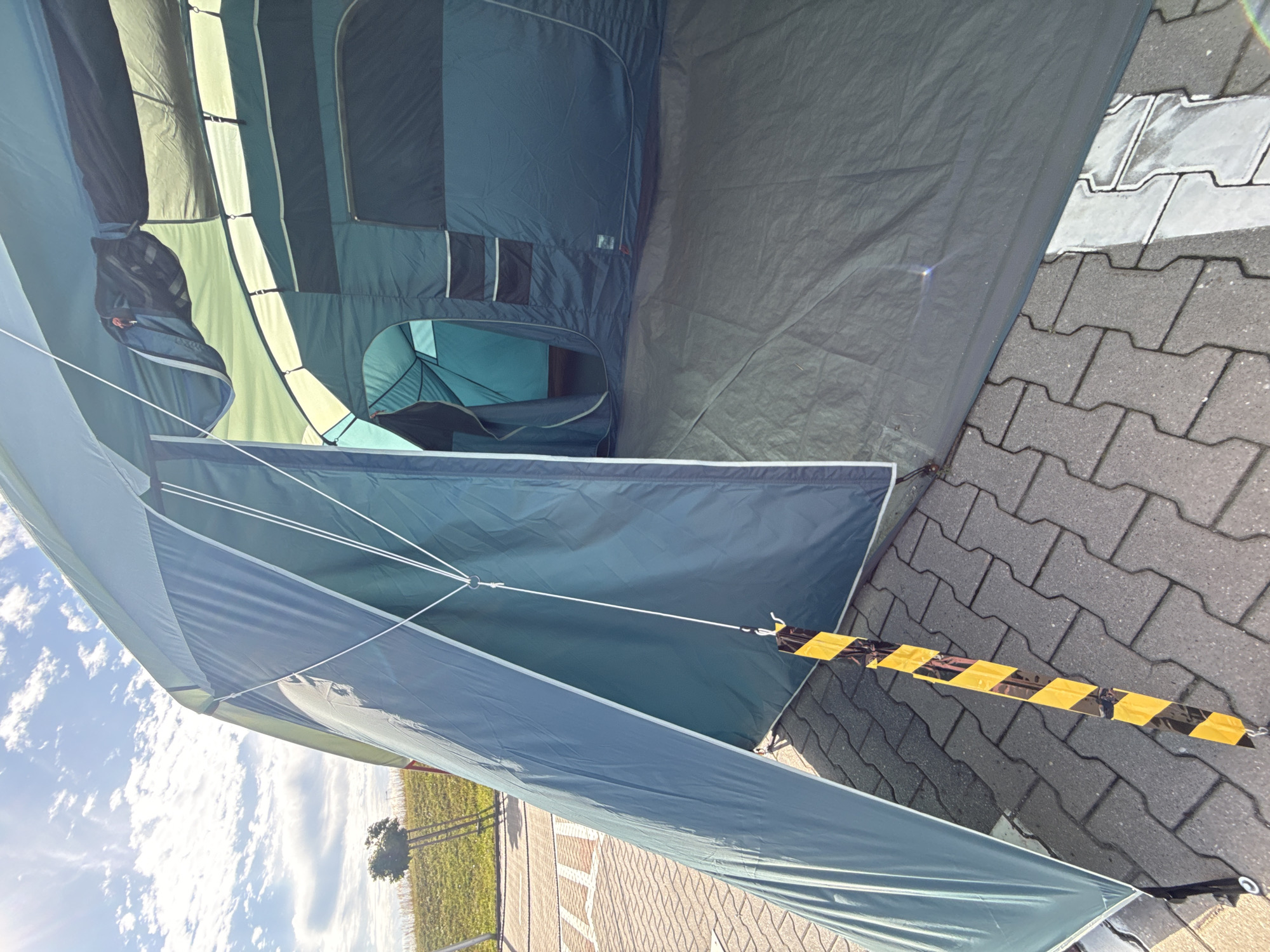 The image shows a large, dark teal camping tent set up on a paved area, likely a parking lot, with interlocking stone pavers visible. The tent has an open entrance, revealing an inner section with multiple compartments and windows. The exterior of the tent is illuminated by sunlight, casting shadows. A black and yellow striped ribbon is tied to a cord extending from the tent. In the background, a grassy slope with a small tree can be seen under a bright blue sky with scattered clouds. The overall scene suggests an outdoor setting, possibly a campsite or an open area near a road. The location is identified as Bielany Wrocławskie, Poland.