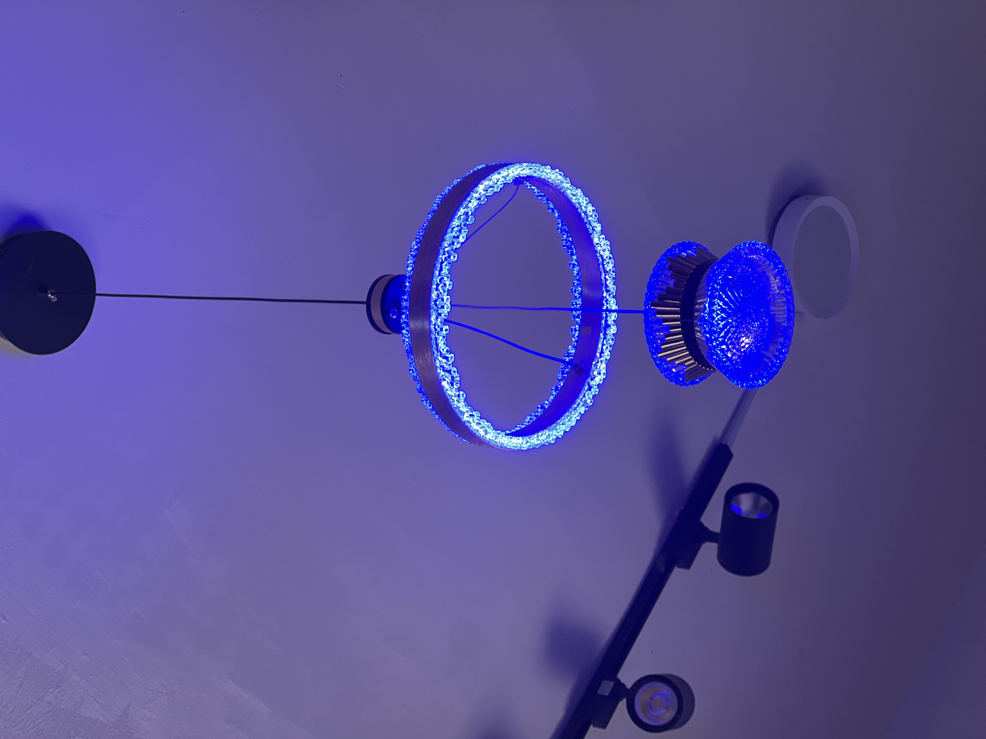 A modern, circular chandelier with blue LED lights hangs from a dark ceiling canopy. The chandelier has a wooden-toned ring adorned with clear crystals or beads that refract the blue light. Below the ring is a smaller, faceted blue glass fixture. To the left, a black track lighting system is mounted on the ceiling, with two black cylinder-shaped spotlights visible. One spotlight points downwards, and another is angled to the left. The ceiling is painted a light, muted purple color and appears to have a textured surface with visible brush strokes. The overall lighting is dim, with the blue lights of the chandelier and spotlights being the primary light sources. There are no people visible in the image.