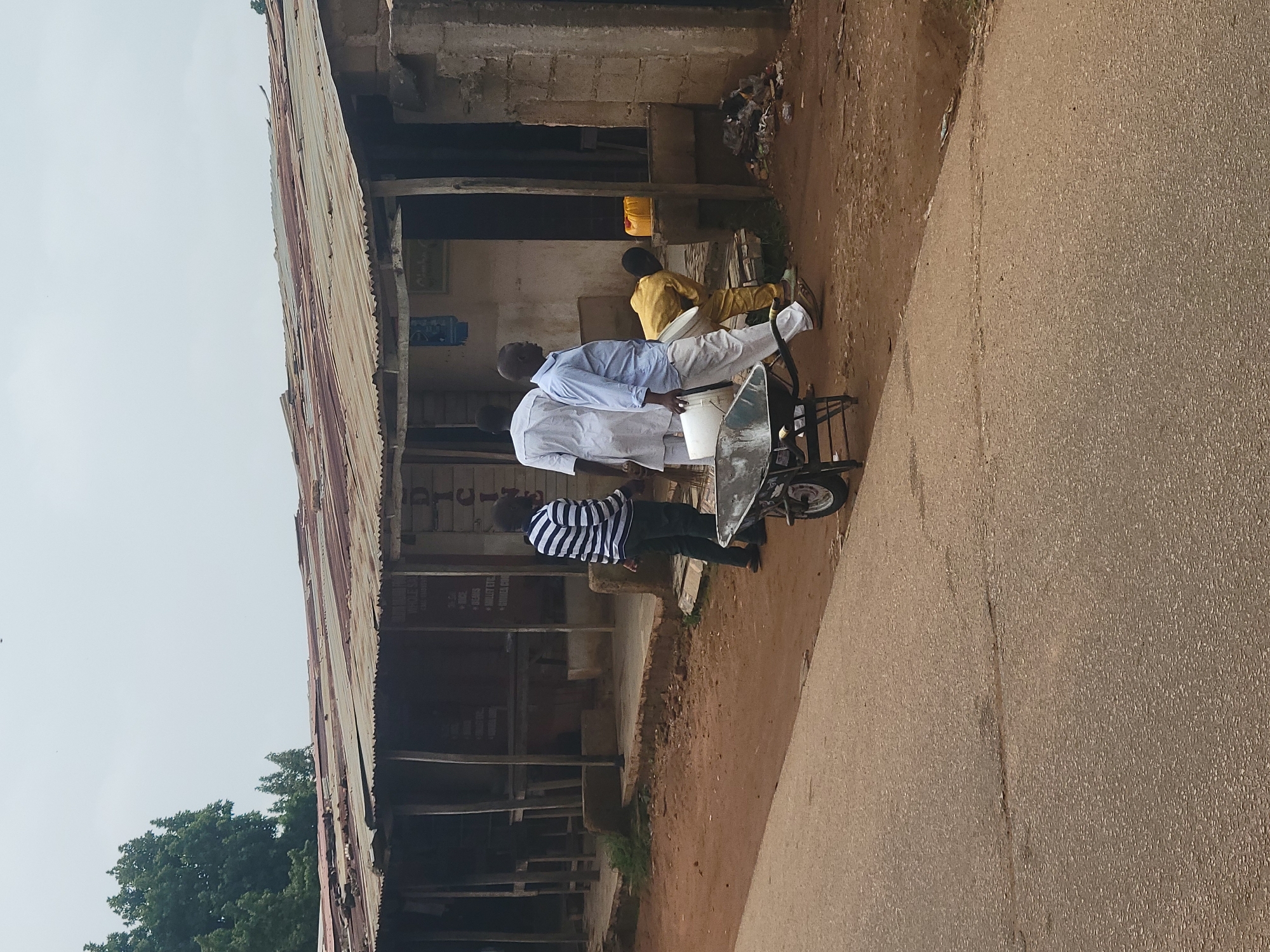 This image depicts a street scene in Jos, Nigeria, featuring a row of weathered, corrugated metal-roofed shops. In the foreground, a paved road runs alongside a dirt path. Several individuals are present in front of the shops.
A person wearing a white robe stands to the left, facing away from the camera. Beside them, a man in a blue shirt and white pants is walking while carrying a white bucket. To the right of him, another individual in a yellow outfit is bending down.
A wheelbarrow is positioned on the dirt path, in front of the man with the blue shirt. The shops themselves appear aged, with visible signs of wear and tear on their roofs and structures. Some items are visible through the open fronts of the shops, suggesting they are places of commerce. The overall environment appears to be a typical urban street in Jos, characterized by its bustling commercial activity and modest infrastructure.