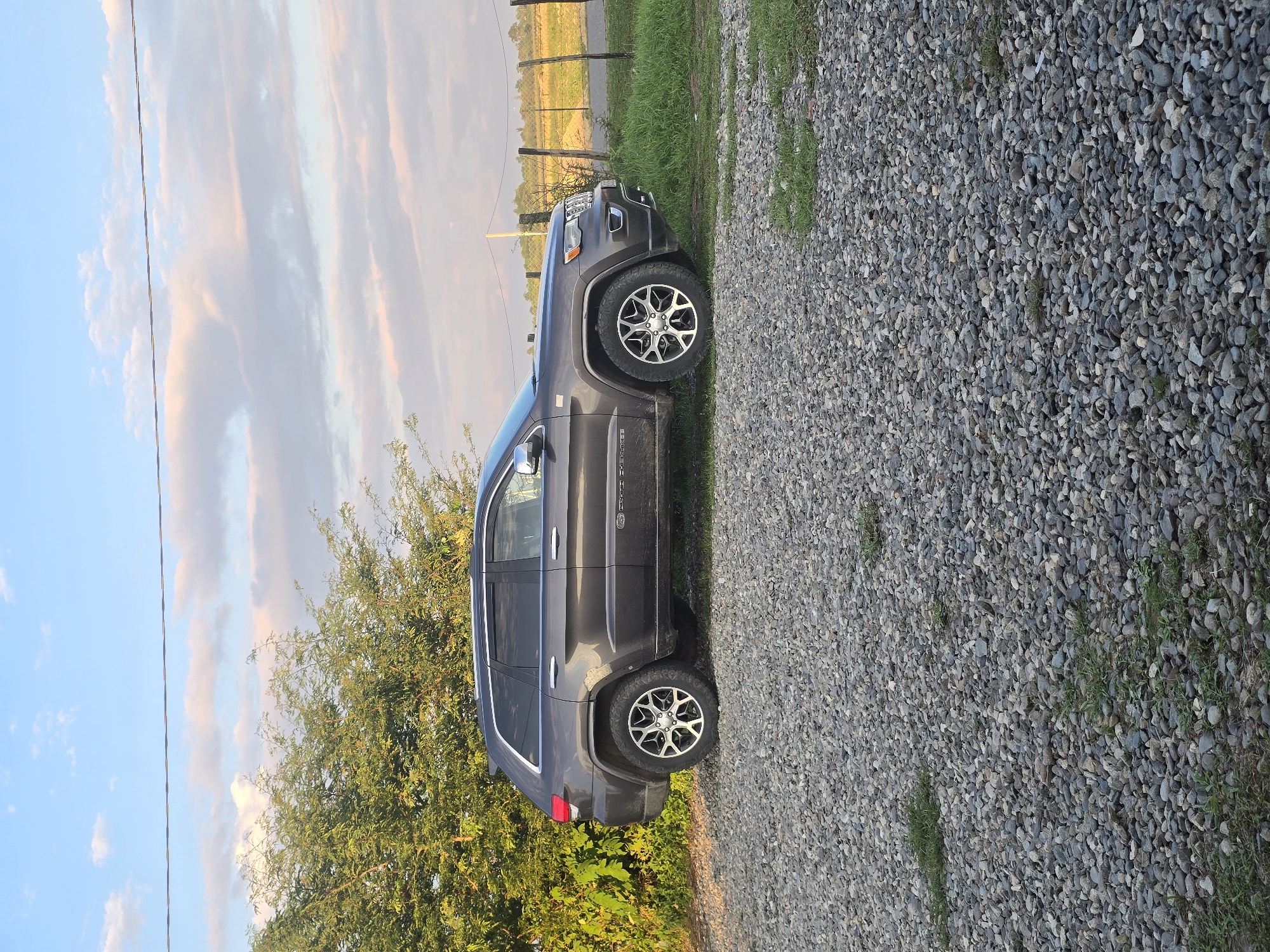 A dark gray Jeep Grand Cherokee is parked on a gravel driveway. The vehicle is facing the right side of the frame, with its rear end slightly closer to the viewer. Lush green trees with vibrant foliage are visible to the left of the Jeep. In the background, a wire fence runs horizontally across the scene, separating a grassy field from a road. The sky above is a soft blue, dotted with wispy white and pink clouds, indicating it is either early morning or late afternoon. The overall environment suggests a rural setting, possibly on the outskirts of Ingiri, Georgia.