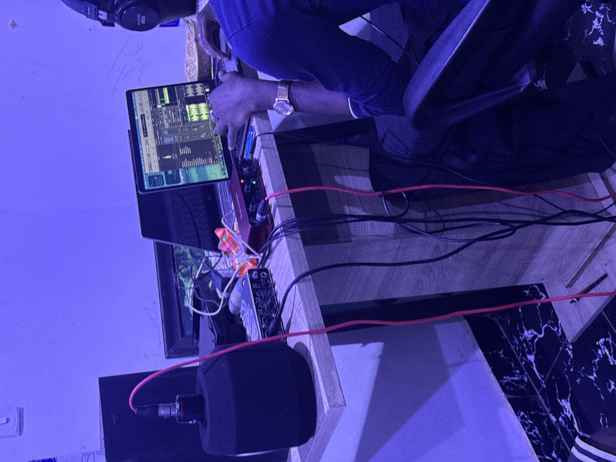 A person is sitting in a chair at a wooden desk in a room with blue lighting. The person is operating a laptop and an audio interface device. A microphone with a pop filter is visible on the desk. Headphones are hanging off the left corner of the desk. Numerous cables are connecting the devices on the desk. The location is possibly in Abuja, Nigeria.