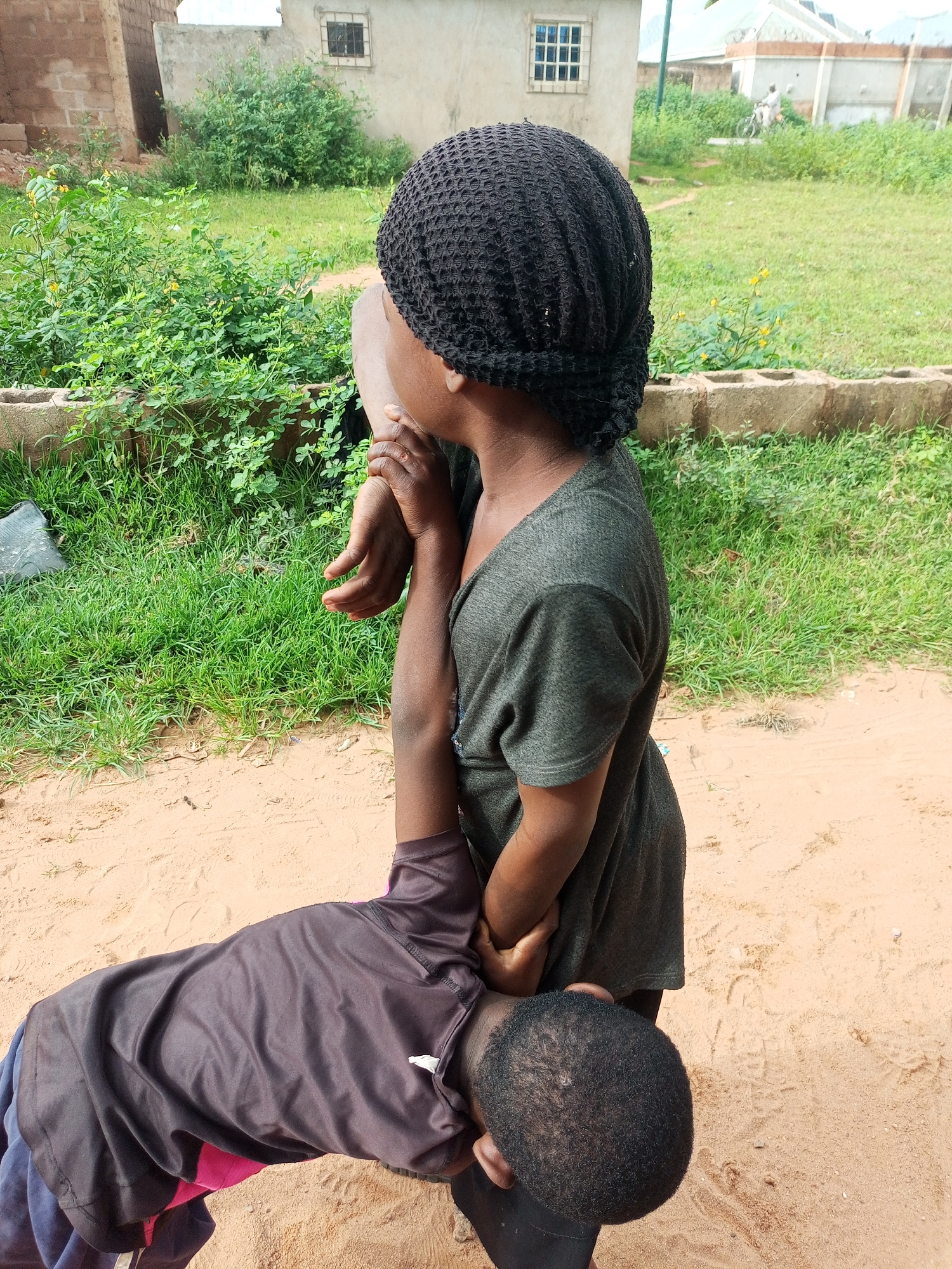 Two children, one standing and one lying prone, interact outdoors in a dusty, sandy area with sparse green vegetation and some simple brick structures in the background. The standing child, wearing a dark textured head covering and a dark green t-shirt, holds the arm of the child lying down. The prone child, wearing a dark t-shirt with pink accents and dark blue pants, is positioned with their head and torso on the ground, facing away from the viewer. The environment suggests a rural or semi-urban setting, potentially a residential area, with evidence of casual construction and an unpaved ground surface. The lighting indicates daytime. The location is identified as Tempure, Nigeria.
