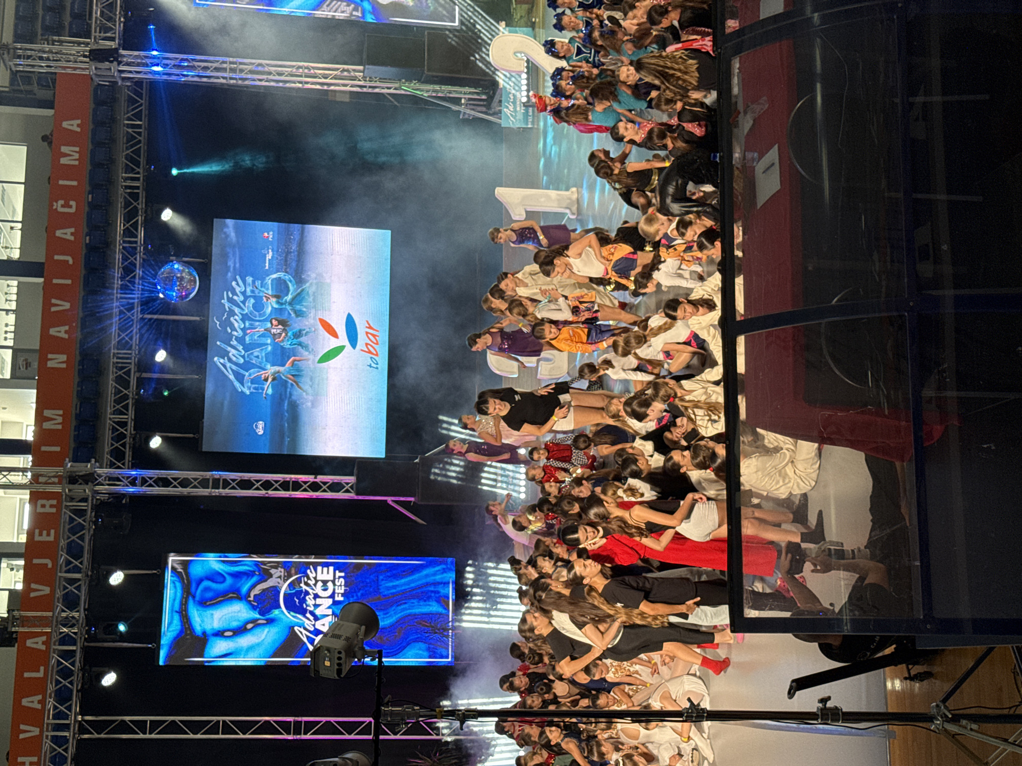 The image captures a large group of young dancers and performers gathered on a stage during what appears to be a dance festival or competition. The stage is set up with large screens displaying graphics related to the event, including the "Adriatic Dance Fest" logo. There are also large decorative numbers, "1" and "2", positioned on the stage. The audience is visible in the foreground, seated in what seems to be an indoor venue. The overall atmosphere suggests a lively and energetic event. The text visible in the background, "HVALA VJERNIM NAVIJAČIMA," translates to "THANKS TO LOYAL FANS" in Croatian and is likely a thank you message to the audience. Based on the "Adriatic Dance Fest" branding and the common practice of hosting such events, it's highly probable this scene is taking place in the city of Bar, Montenegro, a coastal city on the Adriatic Sea known for hosting various cultural and sporting events. The lighting suggests an evening or indoor event.