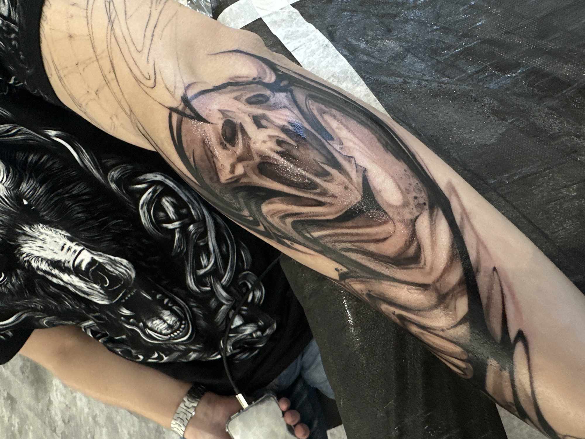 The image shows a person's arm with a partially completed tattoo. The tattoo is a black and grey design, possibly depicting a distorted face or figure. Preliminary sketch outlines are visible on the arm. The person is wearing a black t-shirt with a wolf graphic. A silver watch is on the wrist. The background includes a black textured surface, likely a tattoo studio's workstation cover. The image contains no information that would enable the determination of its location as Moscow, Russia.
