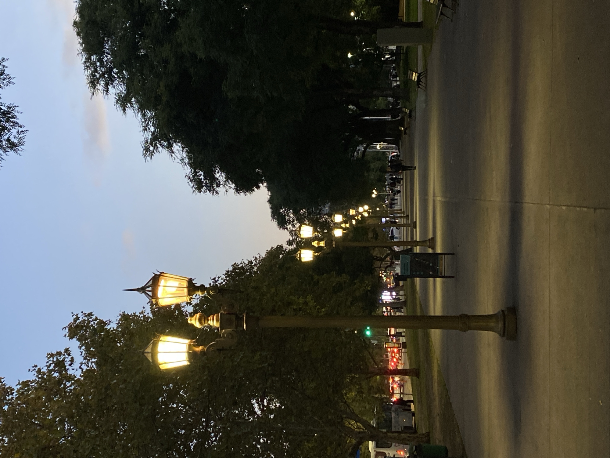 A street in Buenos Aires, Argentina, is lined with trees and ornate streetlights that are illuminated in the evening. There is a concrete or paved path on the left side of the streetlights. In the background, the street extends into the distance, with additional streetlights visible. There are people walking in the distance along the street, and benches are visible along the path. Cars and other vehicles are parked on the side of the street. The sky is a twilight blue.