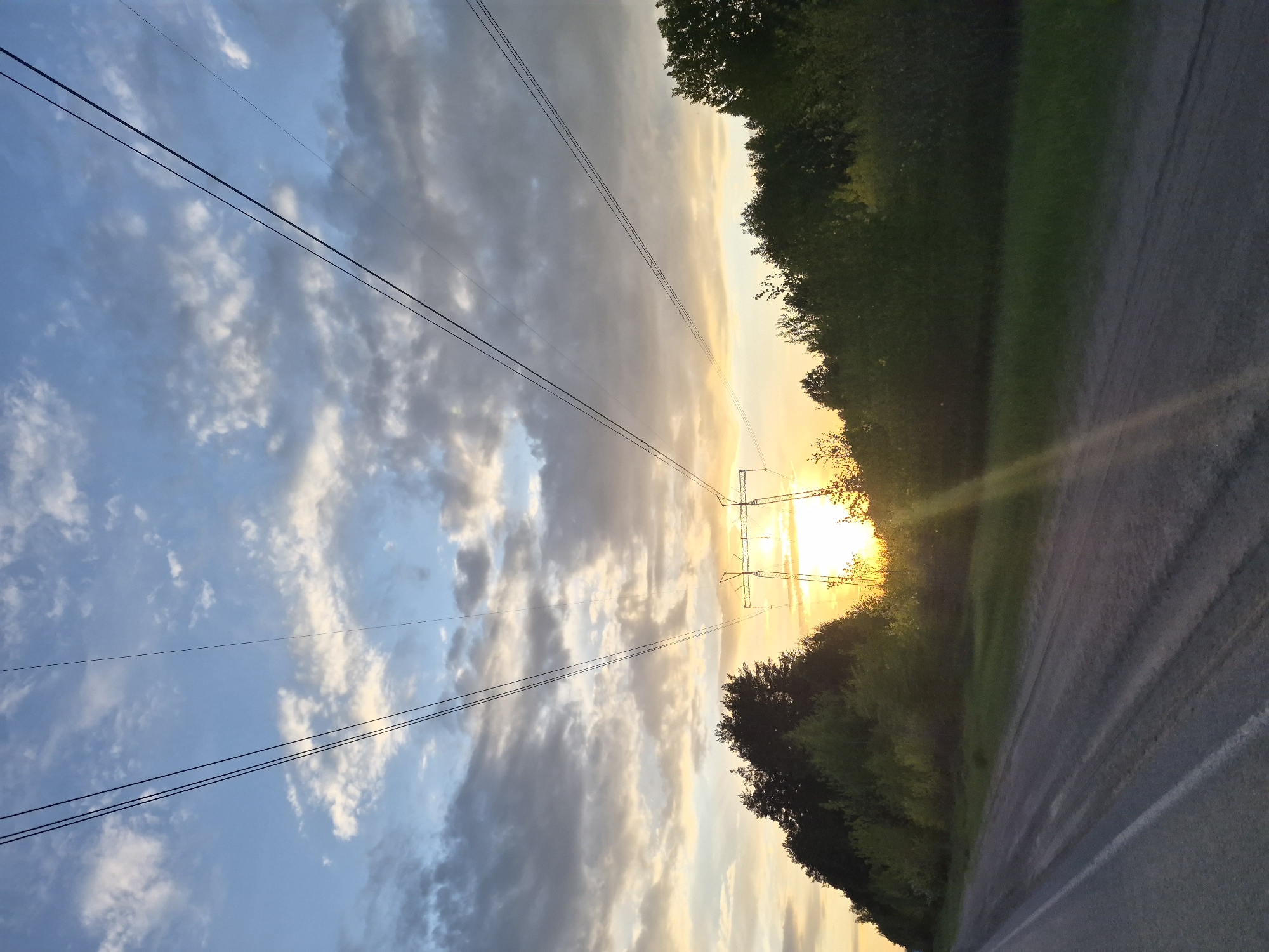 The image shows a landscape at sunset in Noviy, Russia. High-voltage power lines extend across a cloudy sky with a gradient of blue and yellow hues, leading towards a power tower silhouetted against the setting sun. The sun casts a strong glare. A road runs along the right side, adjacent to a line of trees. The picture is slightly tilted.