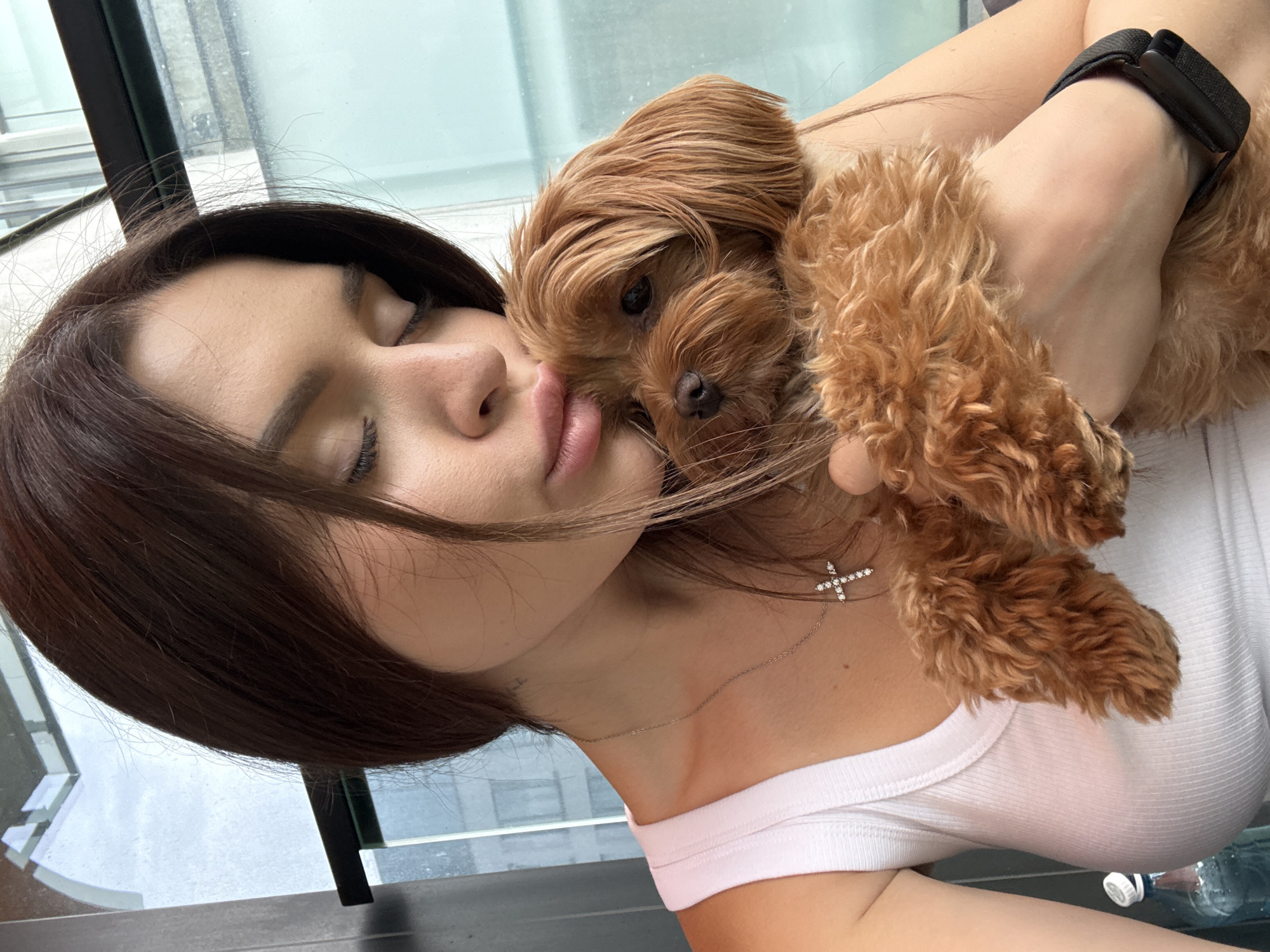 A woman with dark brown hair, wearing a white tank top and a silver cross necklace, is holding a small, light brown dog close to her face. She is leaning her head down towards the dog, with her eyes closed and lips slightly parted, in a pose of affection. The dog, which appears to be a poodle mix, is nestled in her arms, looking forward with its mouth slightly open. A dark watch is visible on the woman's left wrist. The background suggests an indoor setting with large windows, allowing natural light to illuminate the scene. The window frames appear to be black, and beyond them, a blurred urban landscape is visible, possibly indicating a city setting. The lighting is soft and diffused. The image was taken in Prague, Czechia.