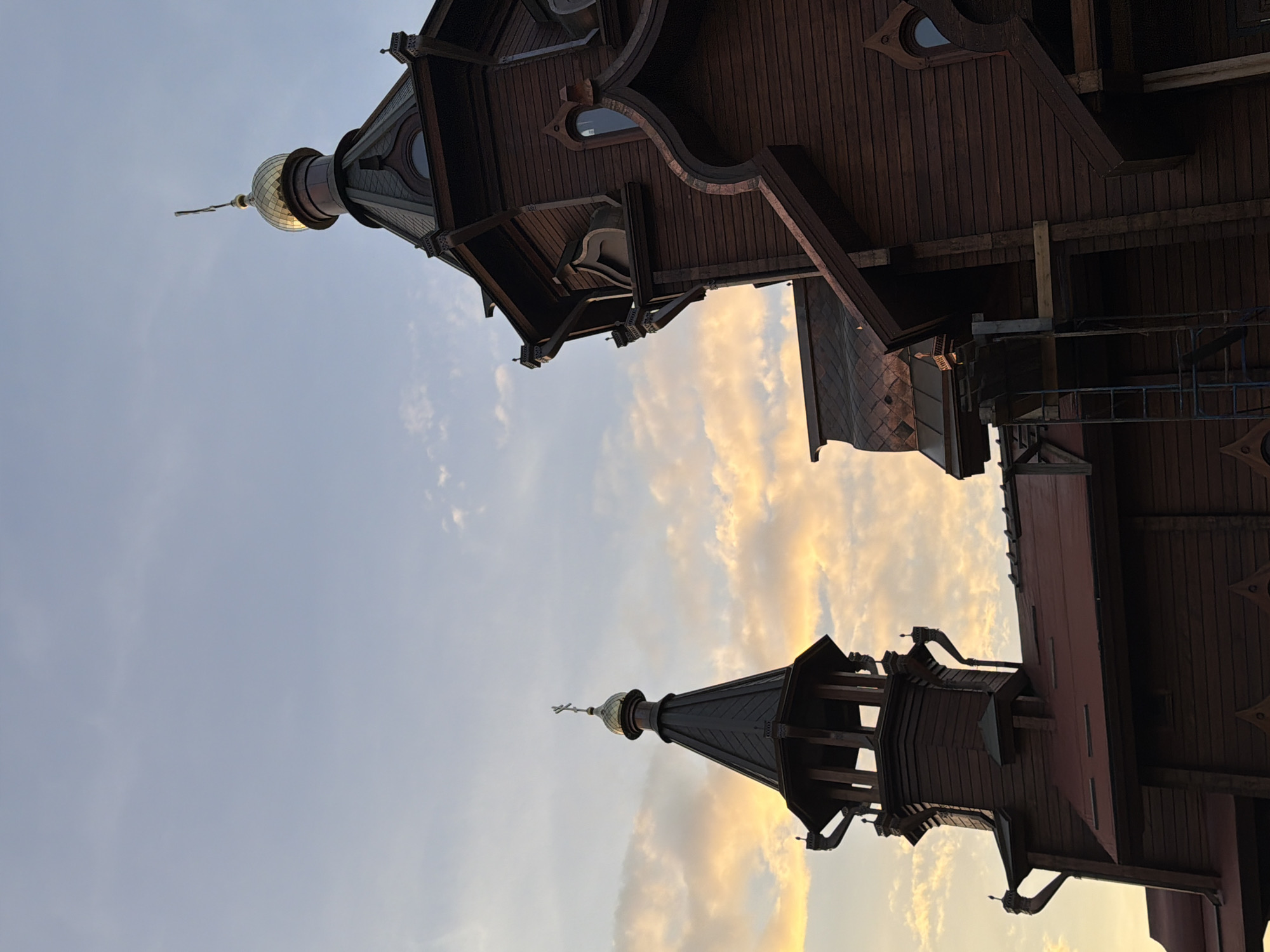 The image features a low-angle view of what appears to be a wooden church or religious building in Krasnoyarsk, Russia, characterized by intricate architectural details and a distinctive style. Two towers are visible with a partially cloudy sky in the background. One of the tower is crowned with a golden dome. The photograph is oriented vertically, emphasizing the upward perspective and the height of the structure.