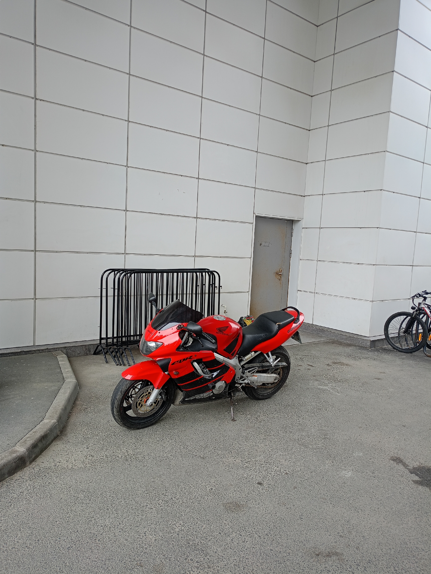 A bright red and black Honda CBR sportbike is parked on an asphalt surface in front of a modern building with a white tiled facade. The motorcycle is angled slightly towards the viewer's left, with its kickstand down. A black metal bike rack is positioned behind the motorcycle. To the right of the motorcycle, partially visible, are two bicycles. A gray metal door is set into the tiled wall behind the bike rack. The scene appears to be an outdoor area in front of a building, possibly a commercial or public space, in an urban environment.