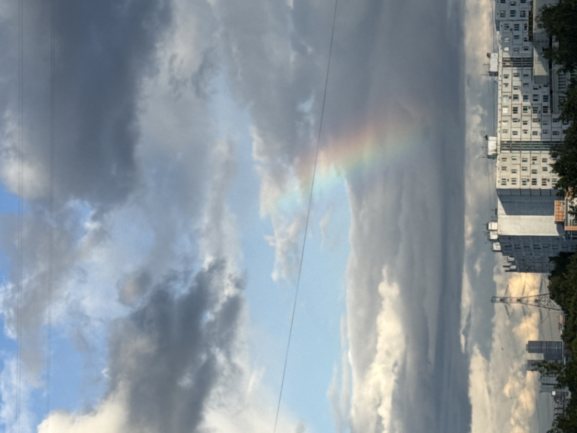 A low-angle, full-shot image captures a fragment of a rainbow arching across a cloudy sky above a city skyline. The sky is dominated by large, dark gray clouds, with patches of brighter blue and white clouds interspersed. A faint rainbow, primarily displaying red, orange, yellow, green, and blue hues, is visible on the left side of the frame. Three thin, dark horizontal lines, likely power lines, traverse the upper portion of the sky. Below the sky, a row of monolithic, white apartment buildings stretches across the lower third of the image. The buildings have numerous rectangular windows and visible rooftop structures. In the background, a large, lattice-style transmission tower stands prominently, with two taller, darker buildings visible to its left. Trees and smaller structures form a dense line along the horizon in front of the apartment buildings. The scene appears to be set in a residential area of a city, possibly during or after a rain shower. The lighting suggests it is daytime, with the sun likely behind the clouds. No people or animals are discernible in the image.