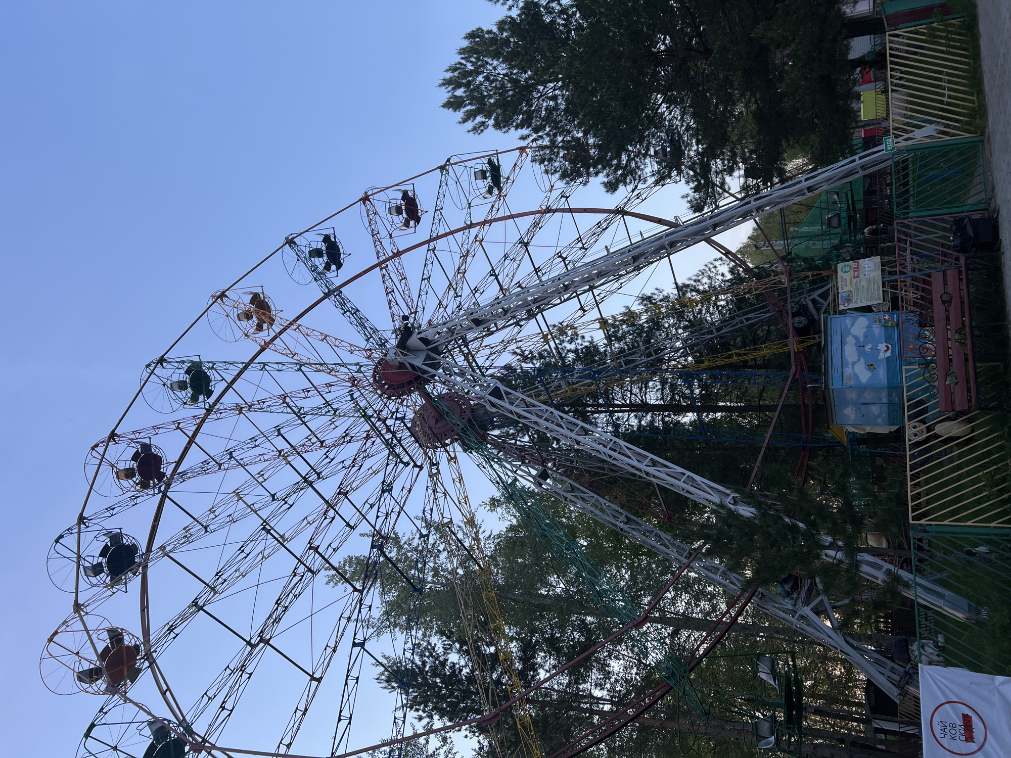 The image shows a Ferris wheel in the city of Chaikovsky, Russia. The wheel is partially filled with passengers. It is surrounded by trees and a blue sky. A fenced area with some structures is to the right of the wheel. A sign with Cyrillic text is visible in the lower right corner.