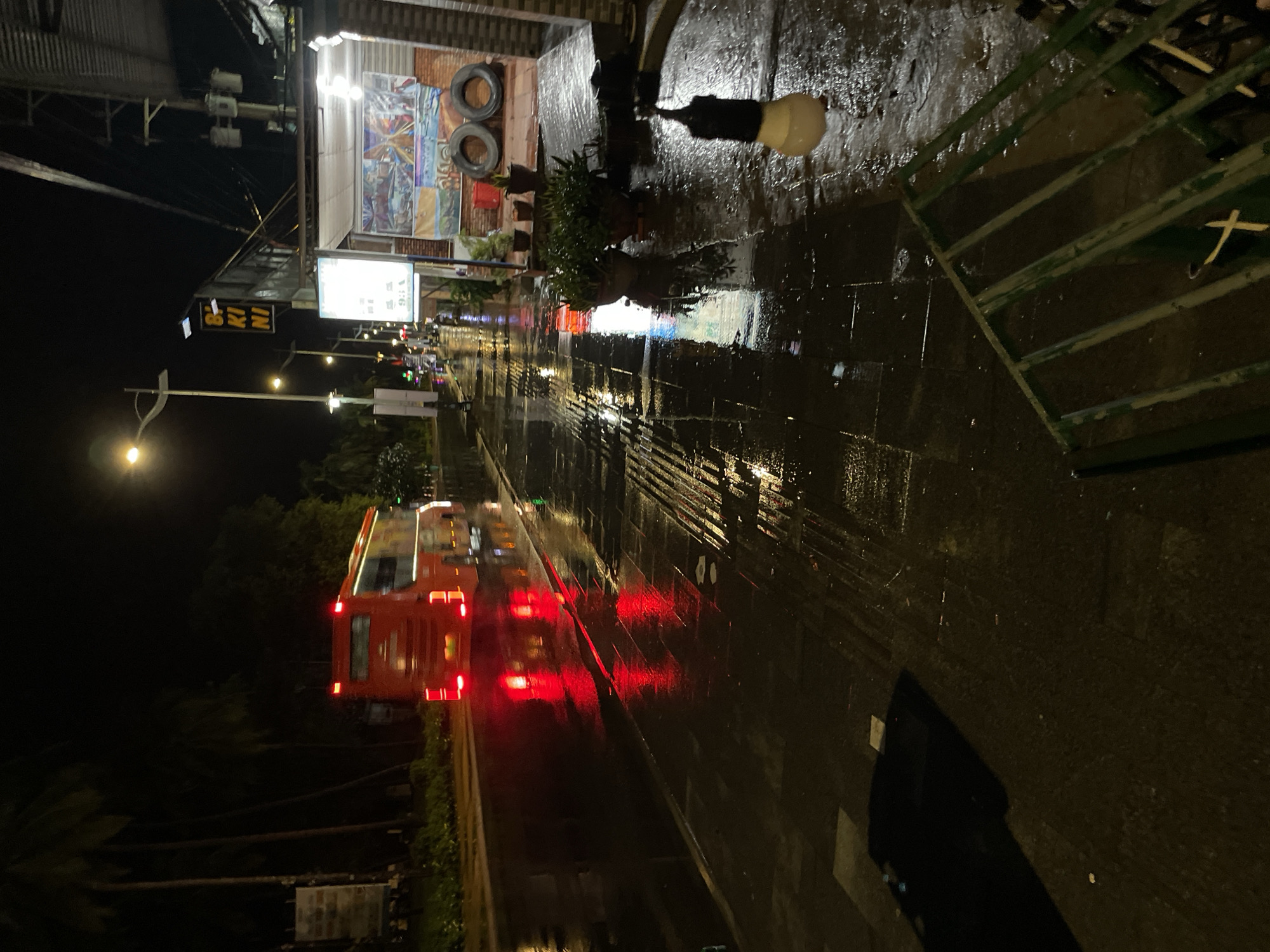 The image depicts a vibrant night scene on a wet street, likely after rainfall, in an urban or semi-urban area, consistent with Phu Thuy Ward, Vietnam. The ground, possibly asphalt or dark tiling, is highly reflective, mirroring the artificial lights and creating luminous streaks.
A prominent red bus or coach, heavily motion-blurred from movement, occupies the left side of the frame, its bright red taillights casting distinct streaks of light on the slick surface. Along the upper side of the street (which would be the right side if the image were rotated to a standard horizontal orientation), several shopfronts or buildings are visible, illuminated by various light sources.
One particular shop features two tires stacked vertically outside and what appears to be a colorful mural or advertisement. Below an awning, a sign with white background and dark text clearly spells out "BIKINI," fragmented into "BI KI NI." Another white sign nearby shows indistinct text and possibly "V:16." Potted plants are arranged along the sidewalk in front of these establishments. A bright, unshaded light bulb hangs down from an awning, contributing to the scene's illumination and reflections. In the upper right corner, a green metal barrier or fence is partially visible. The overall scene conveys the bustling yet atmospheric character of a street at night, with the wet conditions enhancing the visual dynamism of lights and movement.