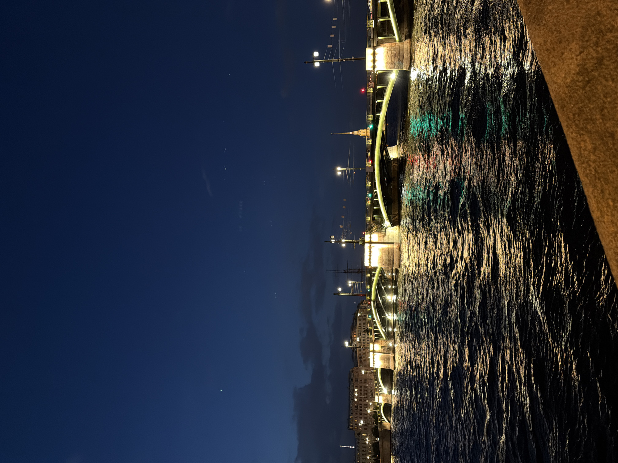 This is a night-time, eye-level, horizontal, medium-shot photograph of the Palace Bridge in Saint Petersburg, Russia, viewed from the Neva River. The sky is a deep, clear blue, with a few faint stars visible. The river in the foreground is dark and choppy, reflecting the lights from the bridge and distant buildings. The Palace Bridge is illuminated by numerous warm-toned lights along its structure, creating a golden glow on the water. The bridge has a distinct arched design, and its stone piers are clearly visible. In the background, several multi-story buildings with lit windows line the riverbank. A tall, slender spire, likely part of a church or administrative building, is visible in the distance above the bridge. There are no people or vehicles visible in the image. The overall impression is one of serene urban beauty at night.