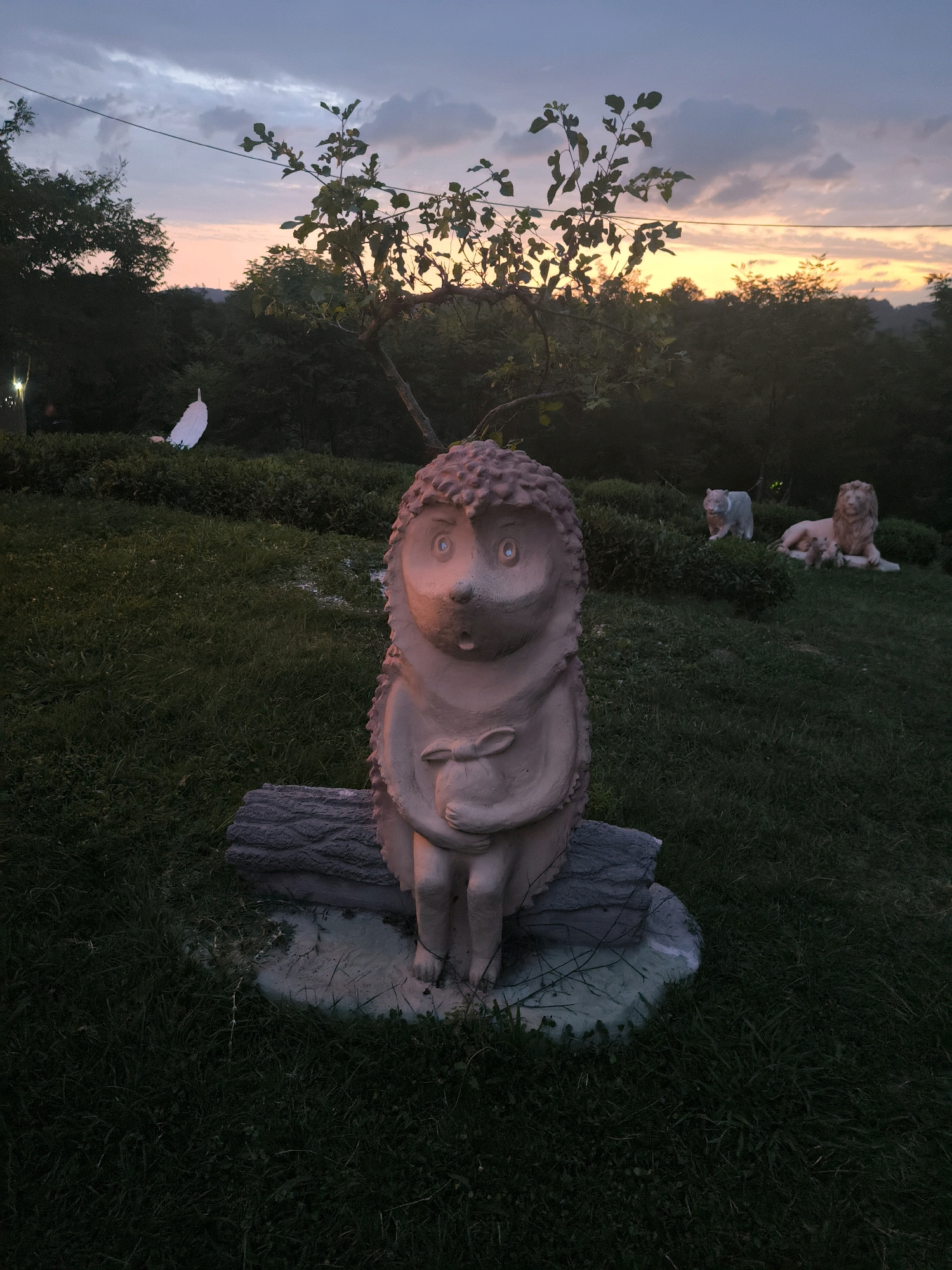 In Kalinovoye Ozero, Russia, a statue of a hedgehog sits on a log in a grassy outdoor area at dusk. The statue is carved from a light-colored material and depicts the hedgehog holding a small object. Behind the hedgehog, two lion statues are visible, one in a seated position and another in a reclining pose. The background consists of dark trees and a sky with dusky clouds and a hint of orange from the setting sun. A single tree with sparse leaves stands to the left of the hedgehog statue. The ground is covered in green grass, and there are some subtle lighting elements, such as a faint light on a distant white object and possibly some illuminated grass in the middle ground.