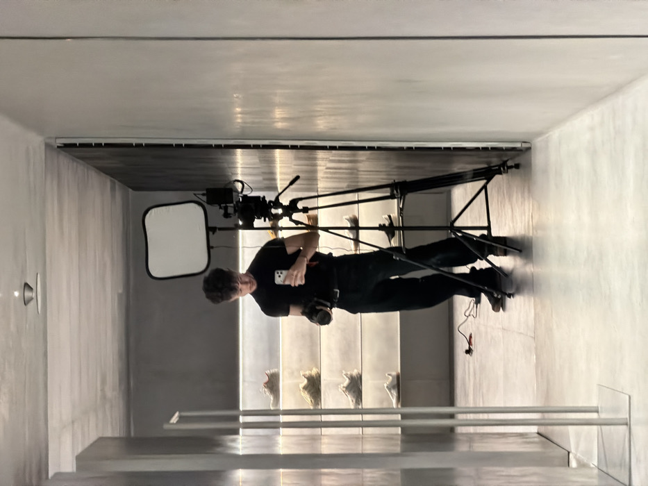 The image captures a male individual taking a mirror selfie inside a modern, minimalist retail store located in Sofia, Bulgaria. The subject, a young adult with short, curly dark hair, wears a black short-sleeved t-shirt, dark trousers, and dark shoes. He is holding a white smartphone in his left hand, angled towards the mirror to capture the reflection, and a professional camera in his right hand, pointed downwards.
To the subject's right, a black professional tripod stands with a camera mounted on it, topped by a rectangular white softbox or LED panel emitting light. A red power cable is visible on the light grey floor near the tripod. The store's interior features light grey walls, with a vertically ribbed textured wall on the left and a smoother wall on the right. Along the back wall, multiple horizontally illuminated shelves display several pairs of modern, chunky sneakers in various neutral tones. A recessed light fixture is visible on the ceiling. On the far left, two vertical silver-colored poles or fixtures are present. The scene suggests a professional photoshoot or video recording either in progress or recently concluded within the store.