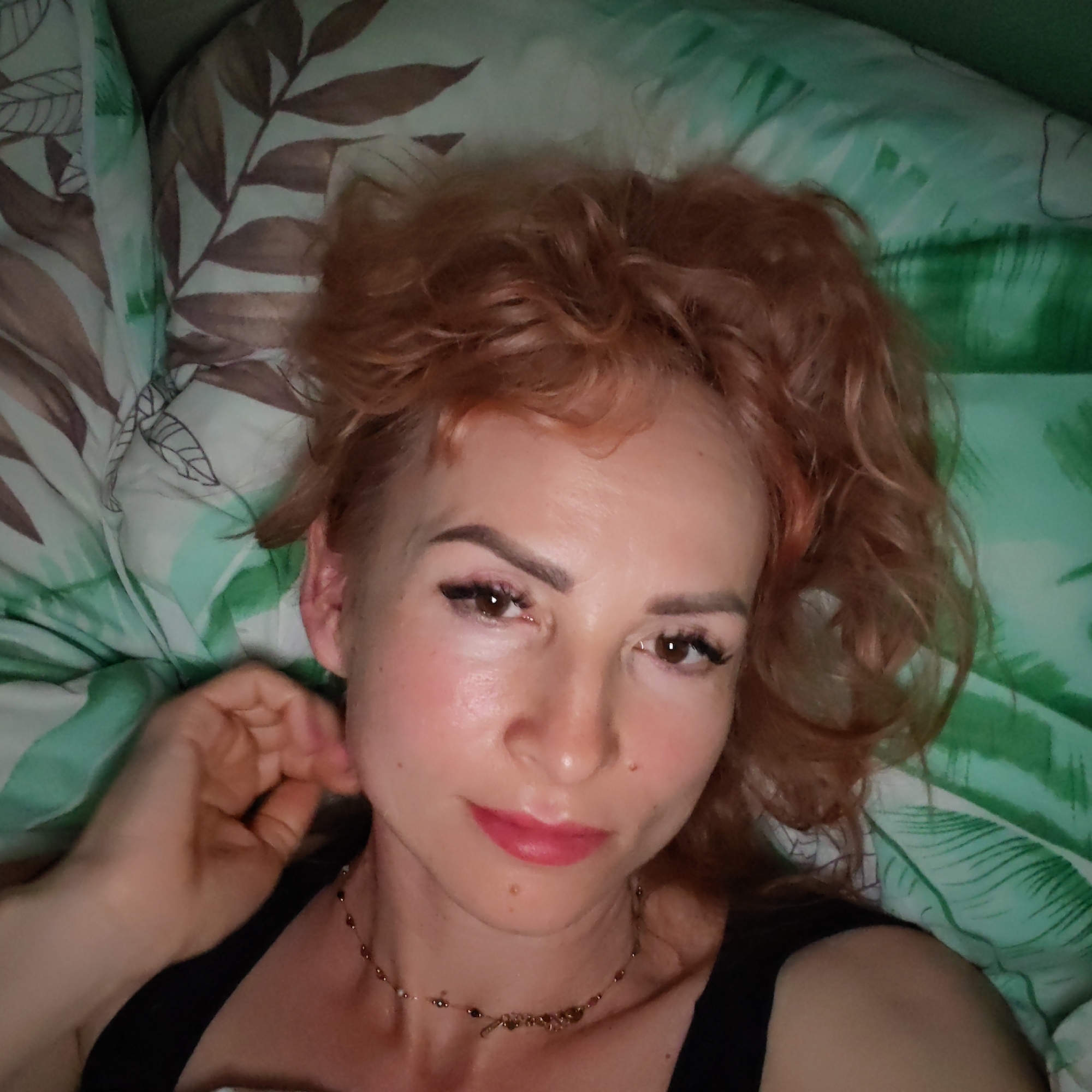 A woman with reddish curly hair is lying on a bed with patterned bedding. She is wearing a black top and a delicate necklace. She has defined eyebrows, eyeliner, and lipstick. Her left hand is near her neck. The bedding has a mix of green and brown leaf patterns. The location is unknown.
