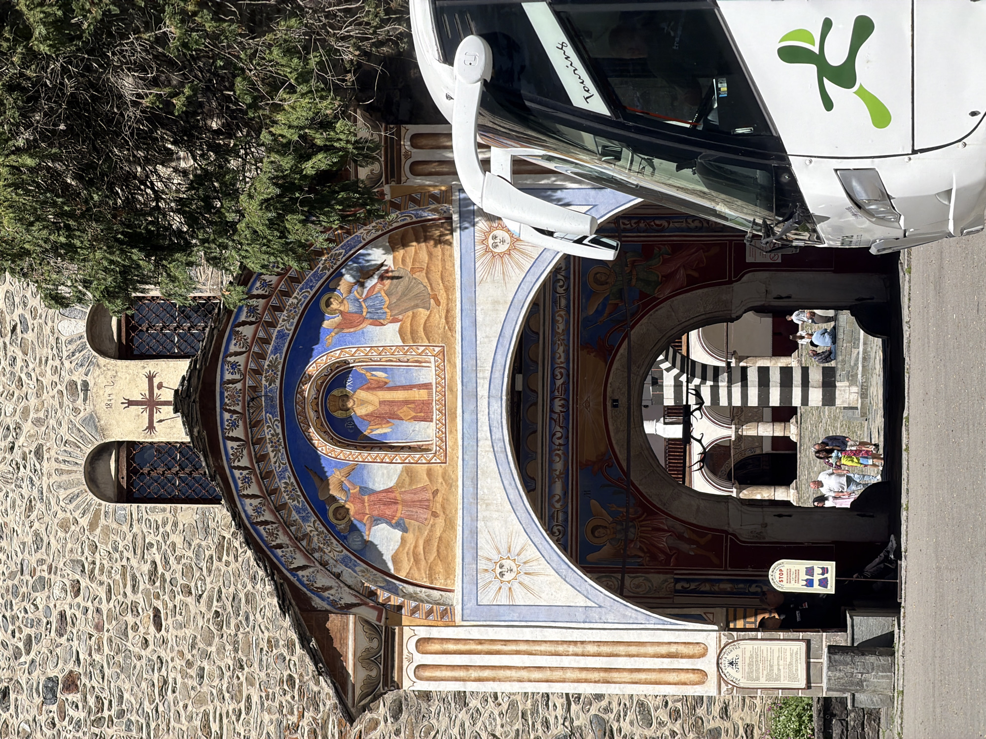 The image depicts a portion of the Rila Monastery in Bulgaria. A stone building with painted murals of religious figures forms a portion of the structure. A white bus with green logo and "Touring" text is partially visible, parked in front of the building. Through an archway, people are visible walking on striped steps. There is a sign on the building facade. A stone wall and a tree with bare branches are also visible.