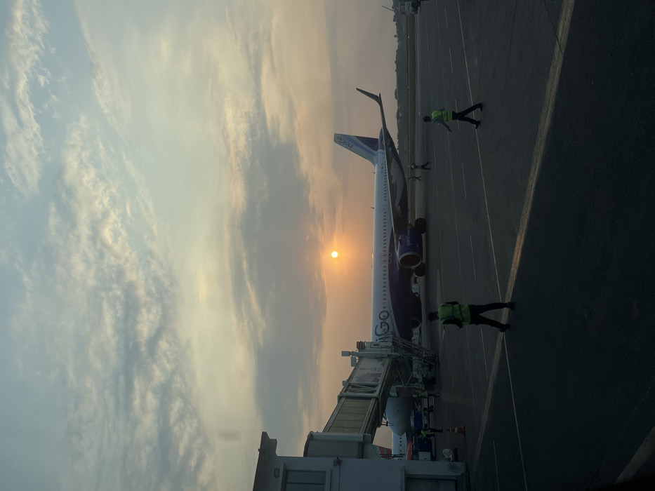 An airport scene unfolds at either sunrise or sunset, characterized by the warm, golden glow of the low sun on the horizon. A blue and white IndiGo aircraft, with "IndiGo" visible on its tail fin and "GO" prominently displayed on its fuselage, is parked on the tarmac. A jet bridge connects to the plane, indicating ongoing or imminent boarding or disembarking activity. Several ground crew members, identifiable by their high-visibility vests, are scattered across the tarmac, attending to their duties near the aircraft. The sky transitions from a vibrant golden-orange around the sun to a softer blue-grey with scattered clouds, creating a scenic backdrop. The dark asphalt of the tarmac contrasts with the illuminated aircraft and sky. The image conveys a calm atmosphere, typical of an airport during these transitional hours, likely in Guwahati, India.