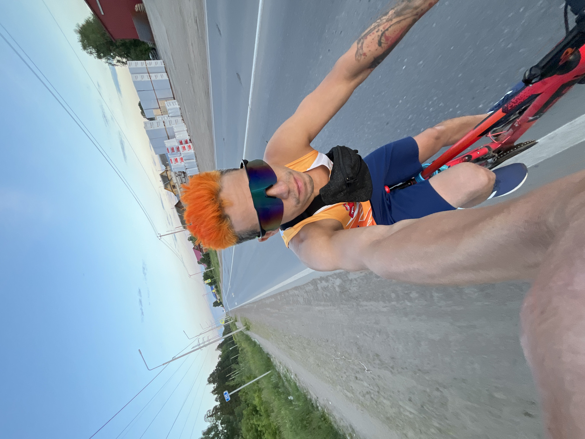 A man with bright orange spiked hair, wearing a sleeveless orange tank top, blue shorts, and futuristic sunglasses, rides a red bicycle on a paved road. He has a black fanny pack around his waist and tattoos on his right arm. The road is lined with a dirt path and green vegetation on the left, and industrial buildings and stacked white materials on the right. The sky is a pale blue with a few wispy clouds. The scene appears to be on the outskirts of the city of Chaikovsky, Russia, likely in an industrial or commercial zone near a residential area. The lighting suggests it is either early morning or late evening.