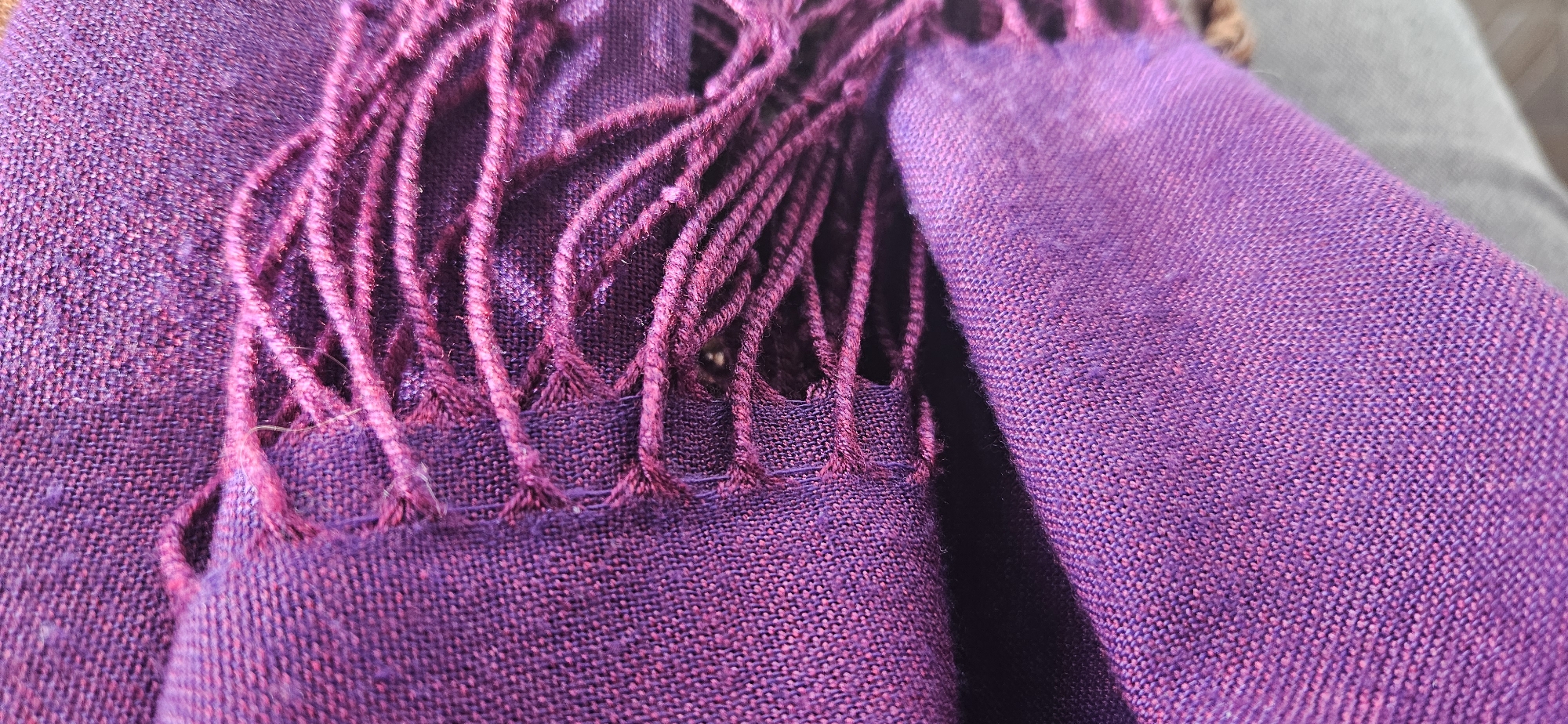 The scene presents a close-up, high-detail view of a purple textile, described as being located in the city of Rila, Bulgaria. The fabric is a woven material, displaying a primary deep purple hue with discernible intermingled reddish-pink threads, creating a slightly mottled or heathered texture. A significant portion of the image is dedicated to the fringed edge of the textile, where individual strands of the purple material are twisted into groups, forming an irregular fringe extending into the frame. The fabric is folded, creating a dark, horizontal crevice running across the middle, separating an upper section with prominent fringe from a lower, smoother section of the textile. Minor loose fibers are visible on the surface. A neutral, light-colored surface is vaguely visible in the background, primarily in the bottom right corner, suggesting the textile is resting on an external object or surface. No people, specific objects other than the textile, or explicit actions are depicted.