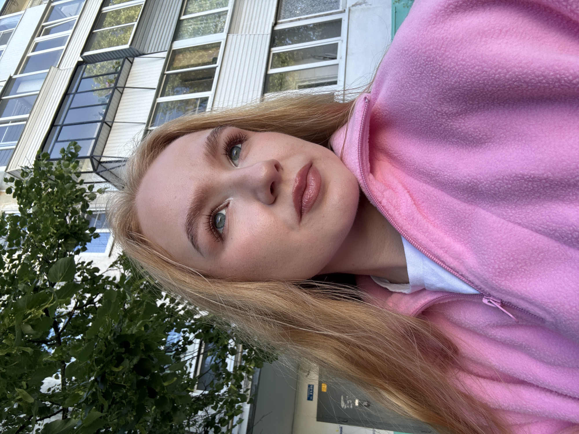 In this close-up, medium shot taken outdoors in the city of Chaikovsky, Russia, a young woman with blonde hair and light blue eyes is captured from the shoulders up. She is wearing a pink zip-up fleece jacket over a white t-shirt and looking off to the right. Her makeup includes eyeshadow and lip gloss. Behind her, a building with multiple windows and balconies and lush green trees are visible. The lighting suggests it is daytime.